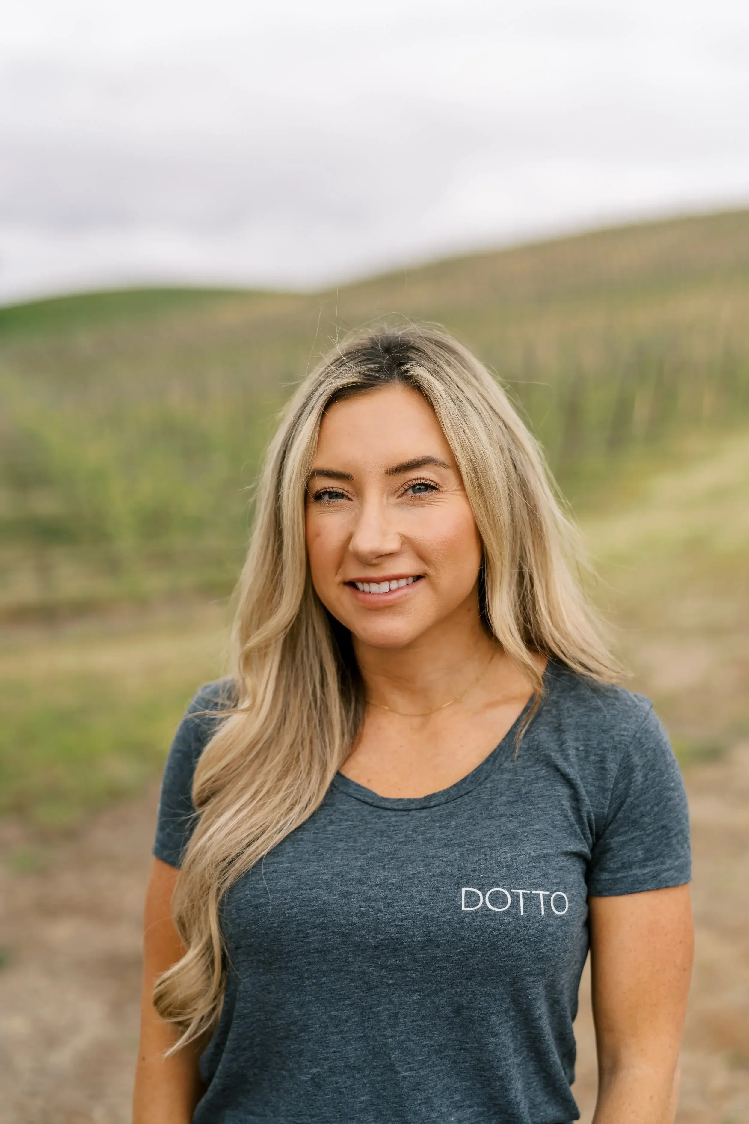 A smiling blonde woman wearing a gray t-shirt with the word 'DOTTO' printed on it, standing outdoors in a vineyard with rolling hills in the background.