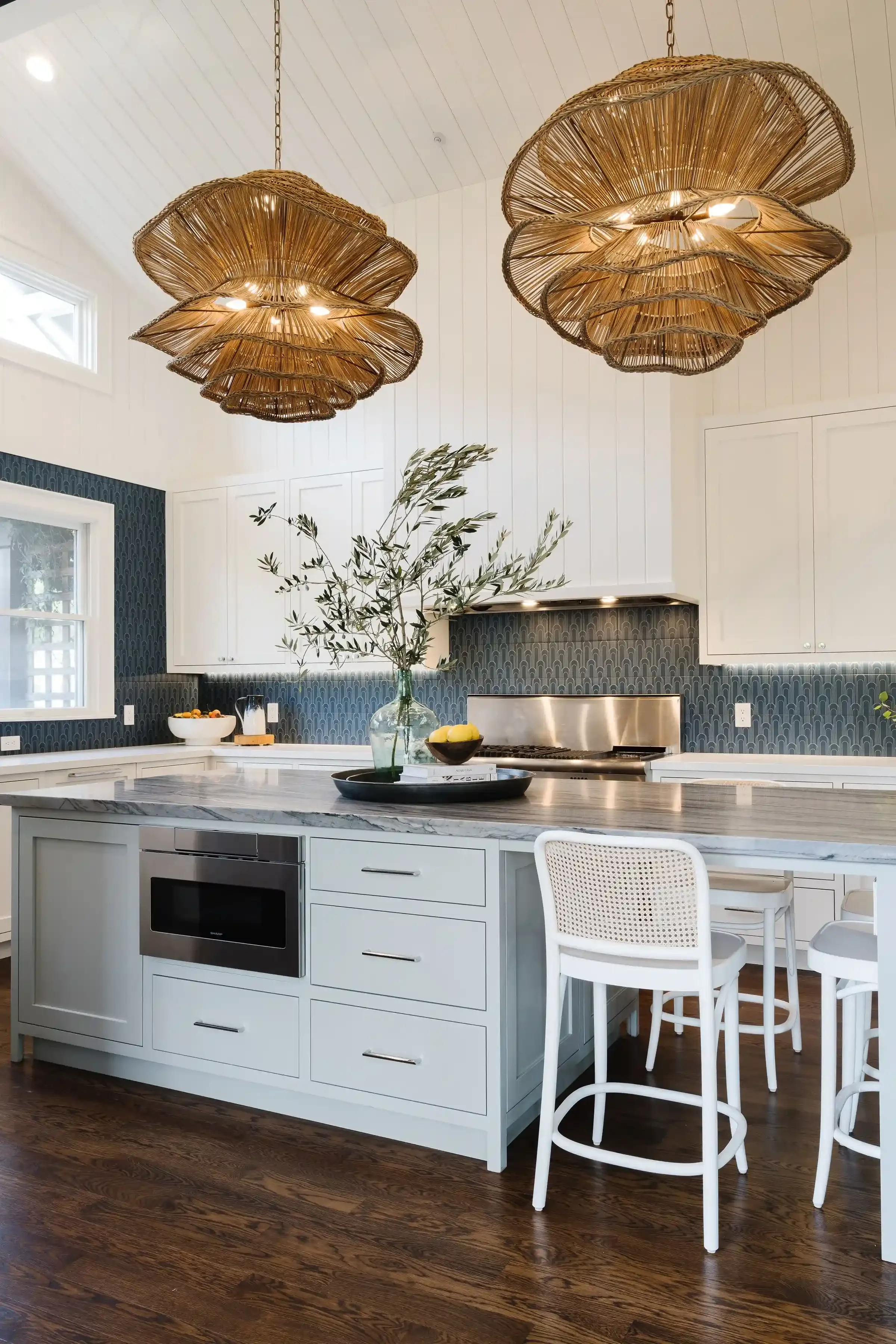 A modern kitchen with white cabinetry, a marble island, dark wood flooring, and unique rattan chandeliers hanging from the ceiling.