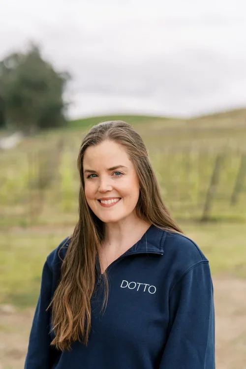 A woman with long brown hair wearing a navy blue jacket with 'DOTTO' embroidered on it, standing outdoors in a vineyard.