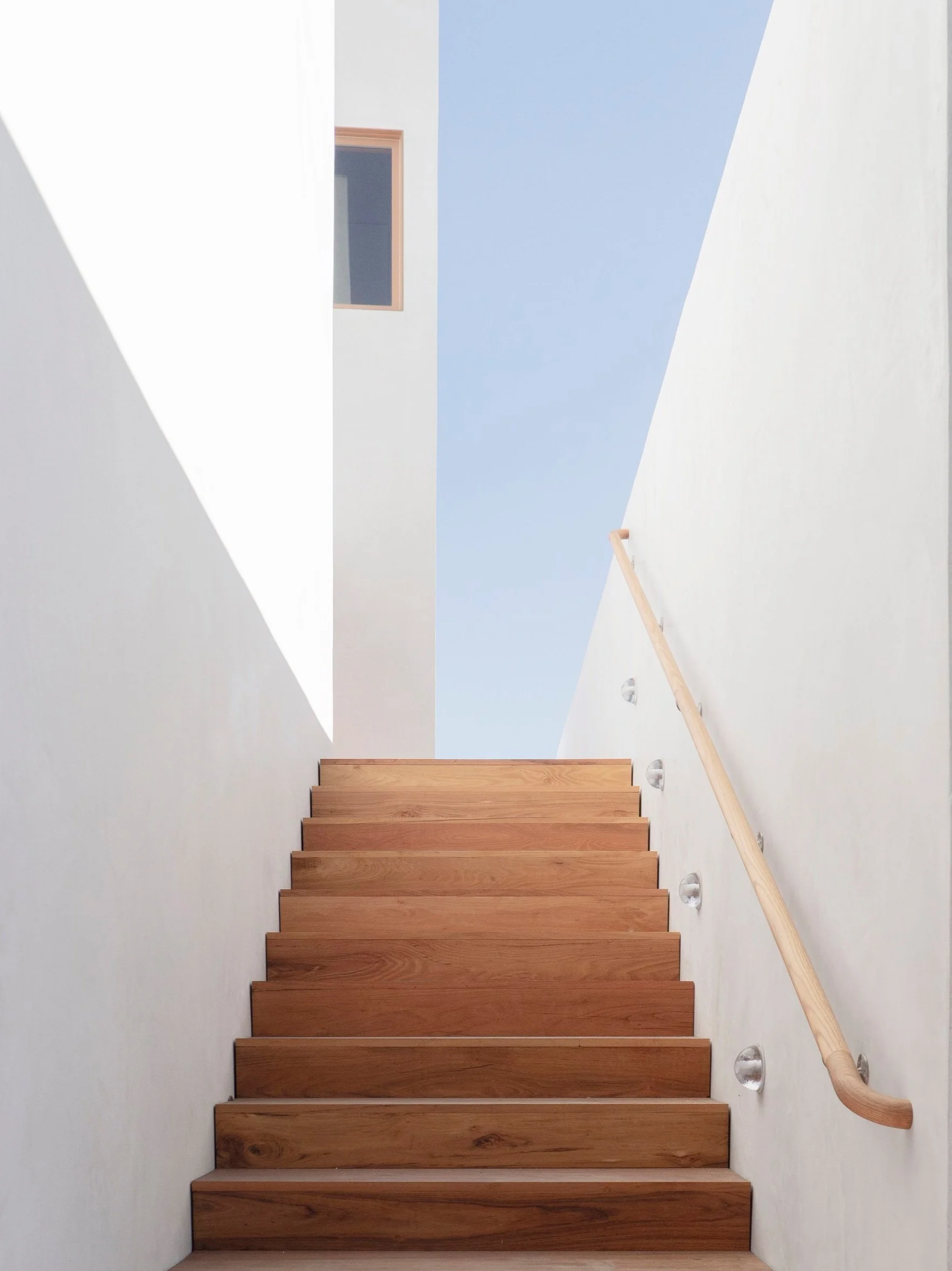 Wooden stairs leading up with a wooden handrail on the right side, white walls on either side, and a clear blue sky visible at the top.