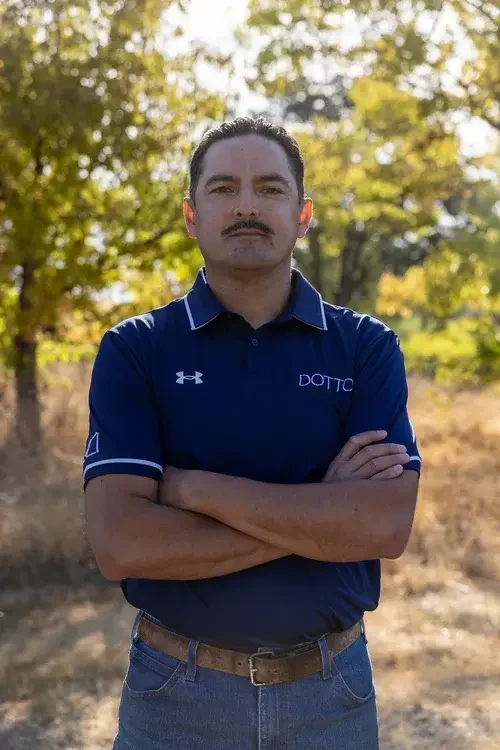 A man with dark hair, a mustache, and light skin standing outdoors in front of trees with yellow and green leaves, crossing his arms, wearing a navy blue polo shirt and jeans.