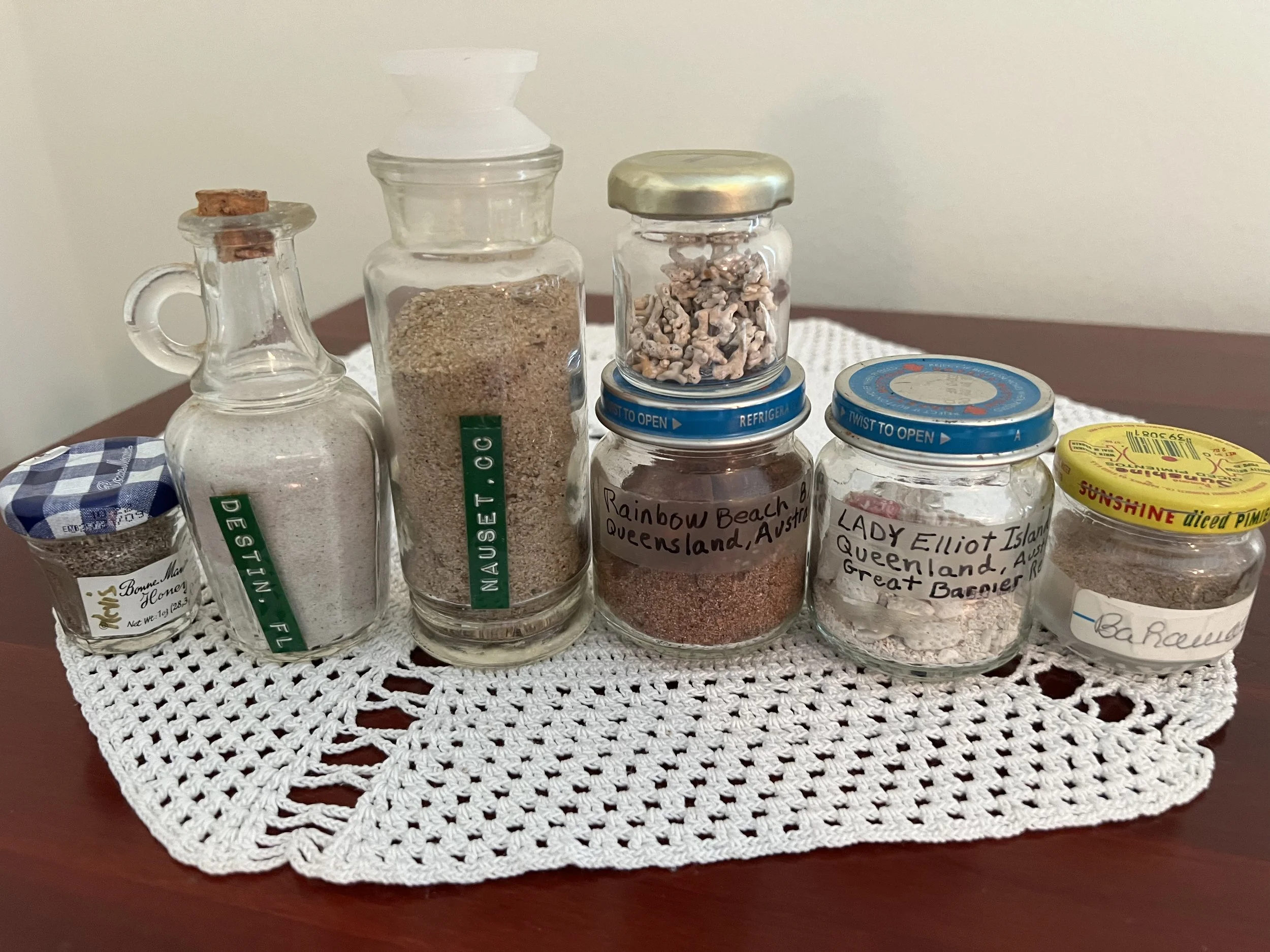 A collection of glass jars containing colored sands and sediments from various locations, including Destin, FL, Nauset, T.C.C., Rainbow Beach, and Lady Elliot Island, arranged on a crocheted doily on a wooden surface.
