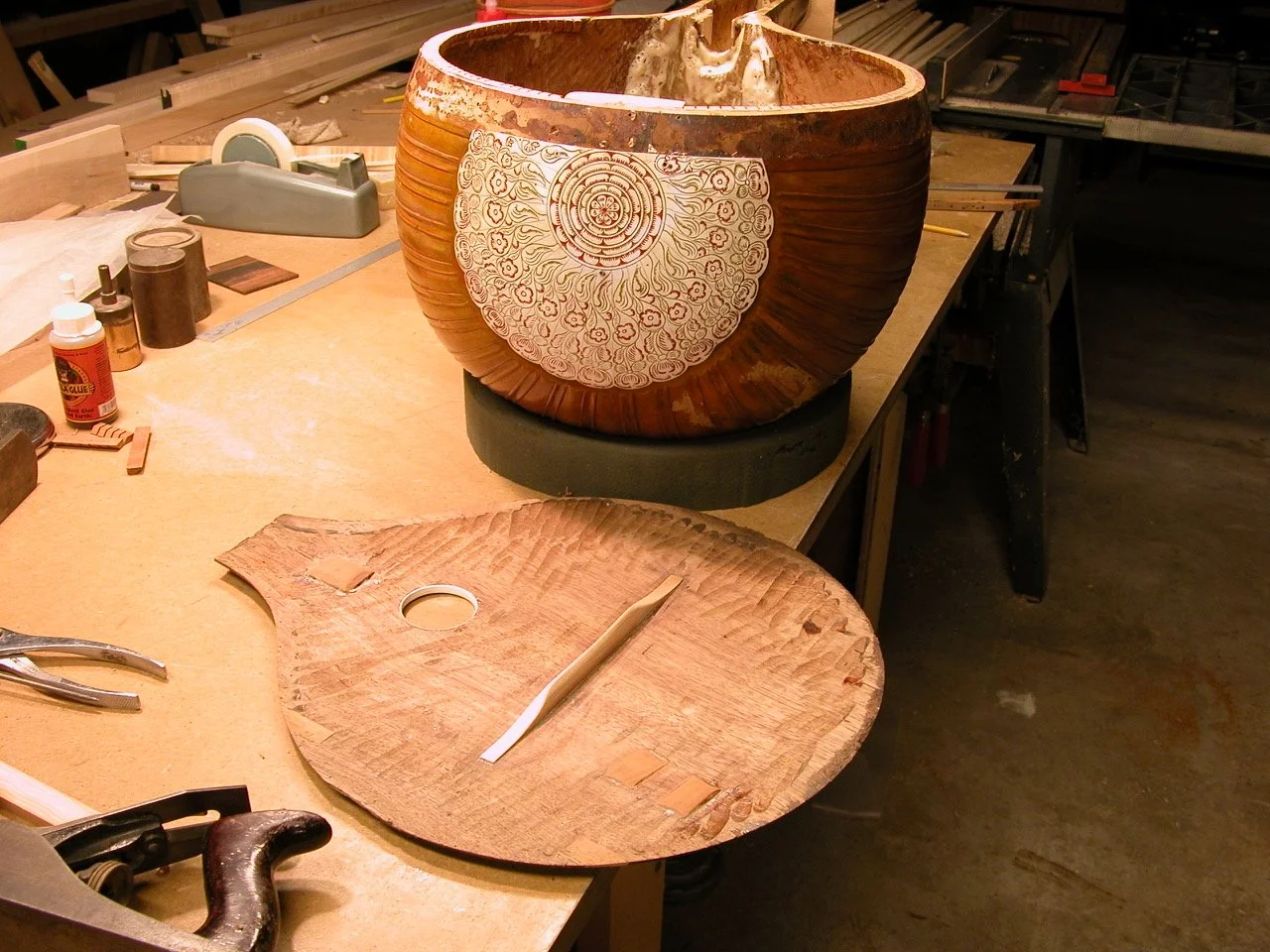 Wooden drum under construction on a workbench, decorated with a white patterned design, surrounded by woodworking tools and materials.