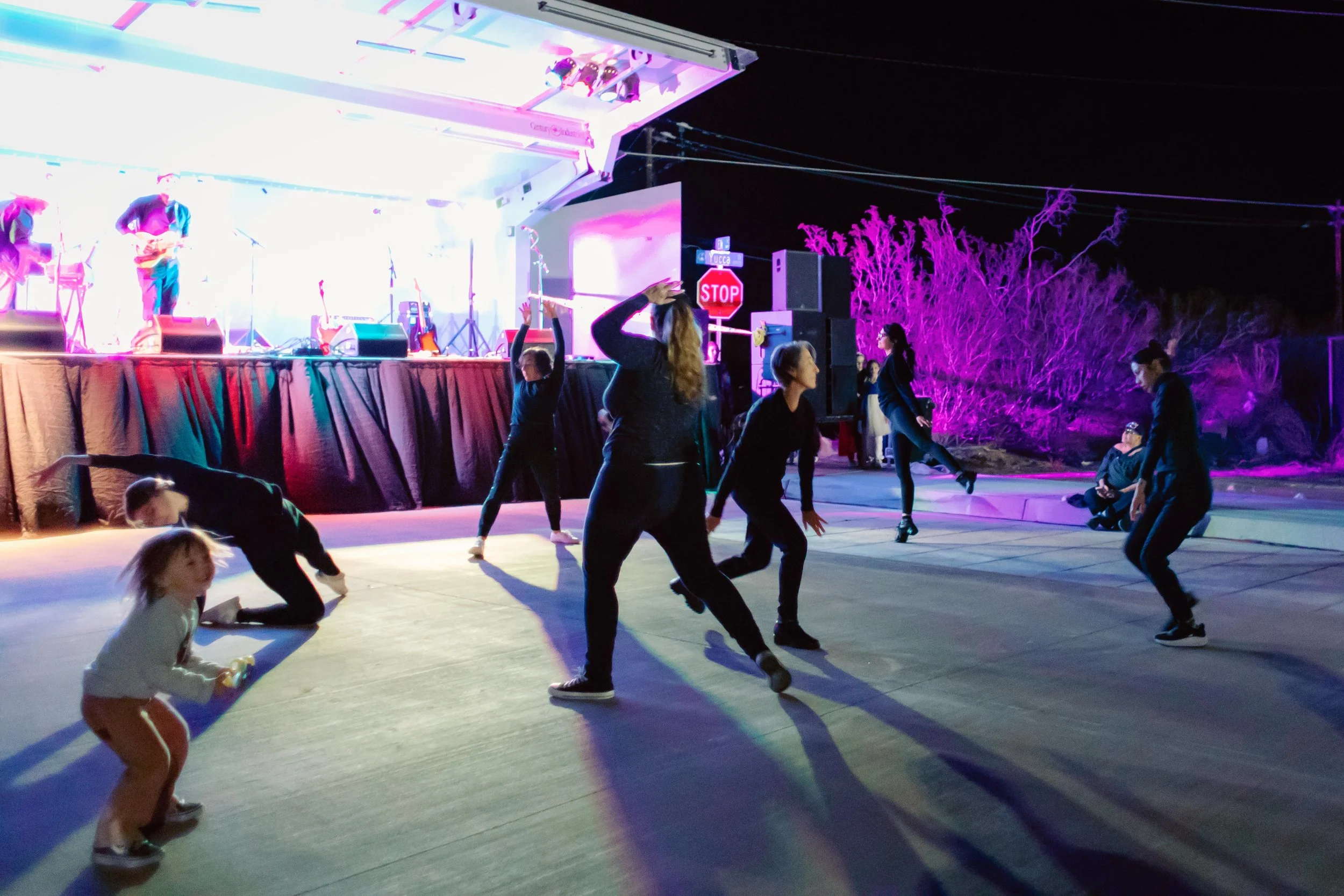 Photo showing dancers outdoors performing on a street with musicians onstage behind them.