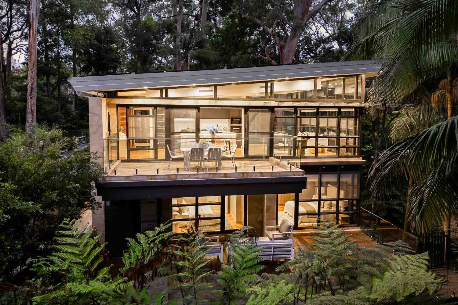 Modern multi-story house with large glass windows and a balcony, surrounded by lush trees.