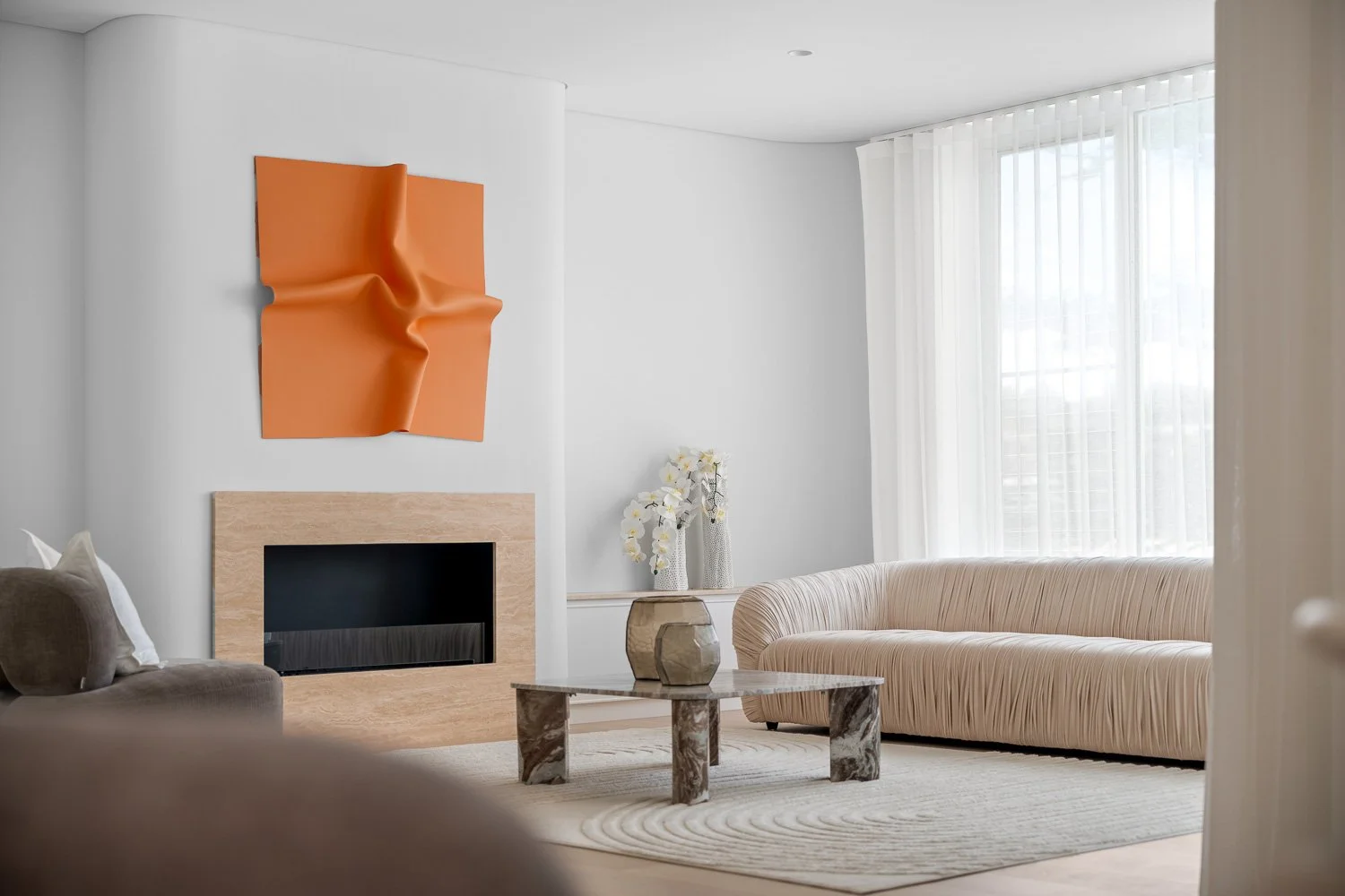 Modern living room with beige sofa, marble coffee table, white walls, orange wall art, fireplace, and large window with white curtains.