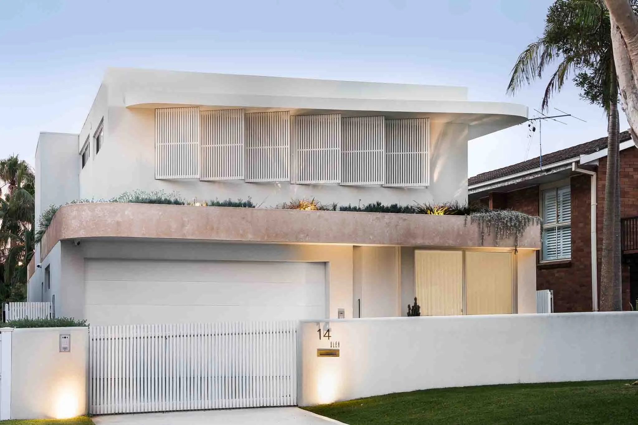 Modern white house with clean architectural lines, a large garage, a small front garden, and horizontal wood slat privacy screens on the upper floor, surrounded by mature palm trees.
