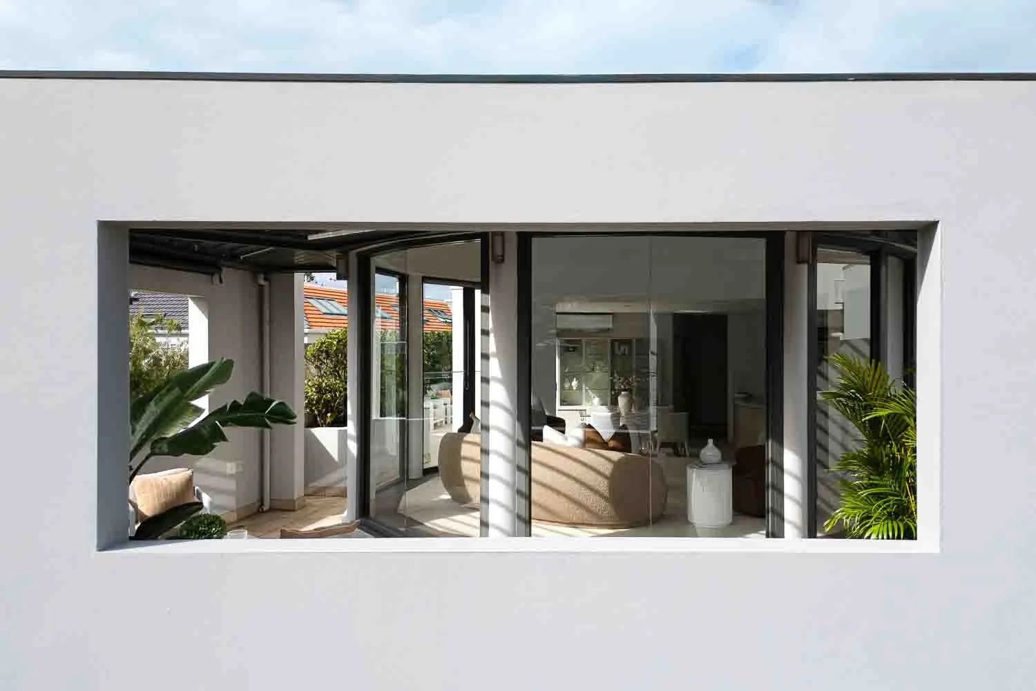 View through a rectangular window showing a modern living room with beige sofas, potted plants, and large glass doors leading to an outdoor balcony.