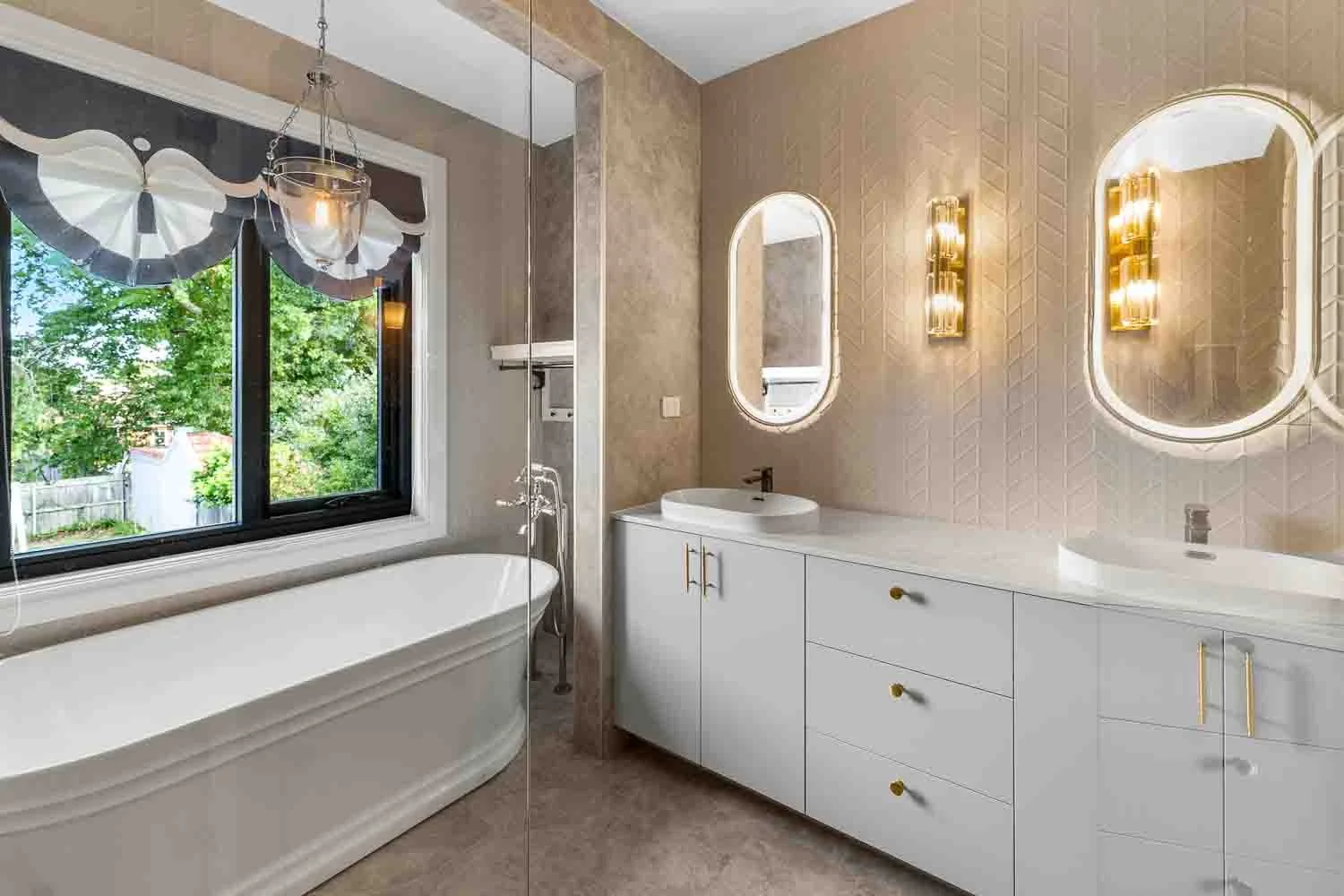 Modern bathroom with a white bathtub near a window with a view of green trees, and a white vanity with two oval mirrors and gold light fixtures.