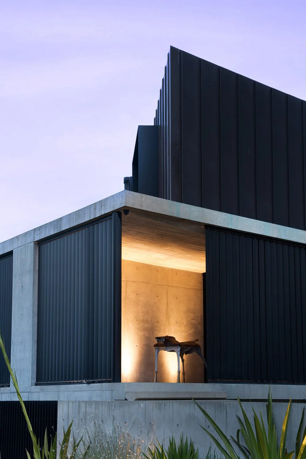 Modern building with black metal siding, concrete accents, and an open courtyard illuminated with warm lighting, showing a sculpture inside.