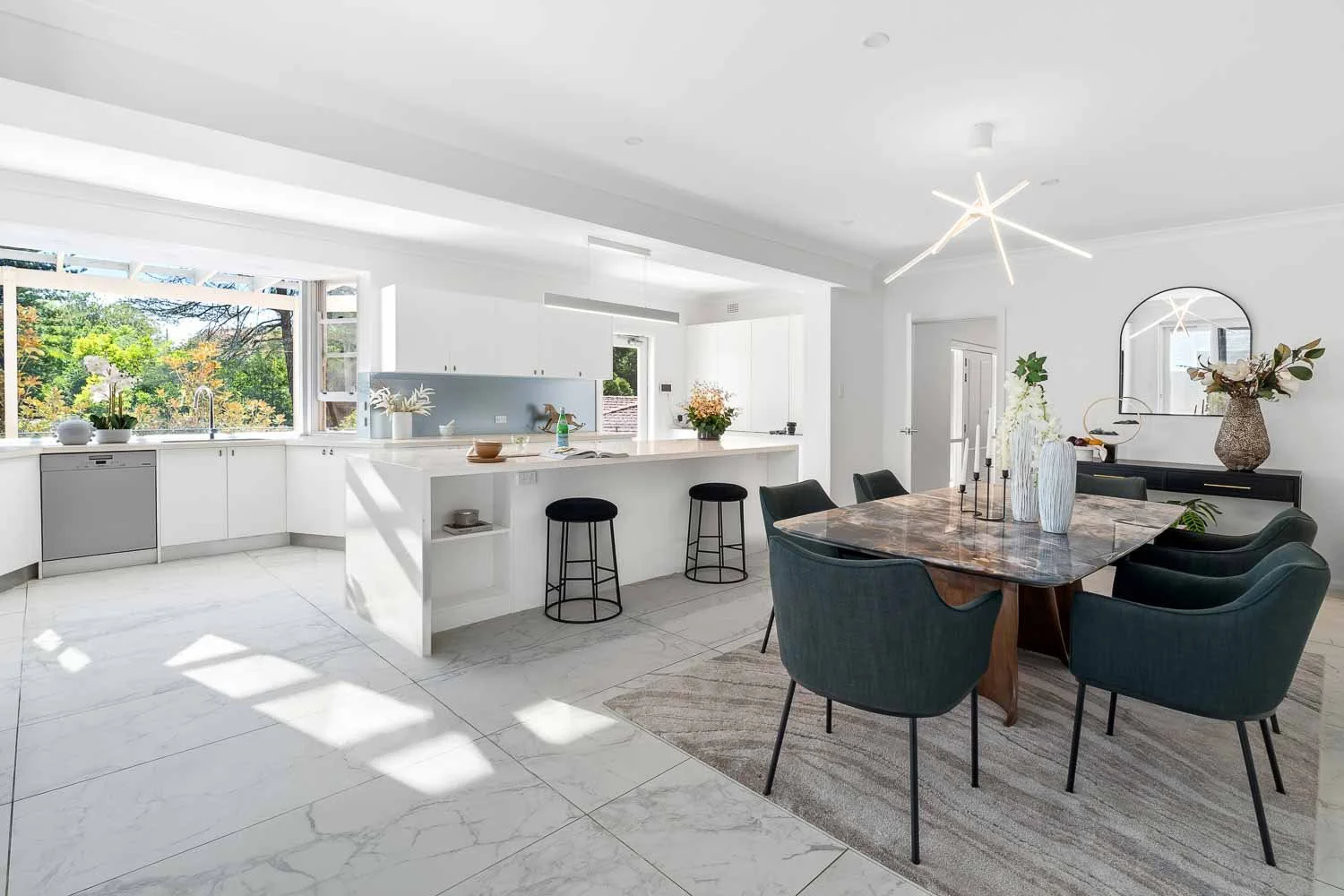 Modern open-plan kitchen and dining area with white cabinets, marble flooring, a large window showing greenery, a marble dining table with six green chairs, and decorative vases and plants.