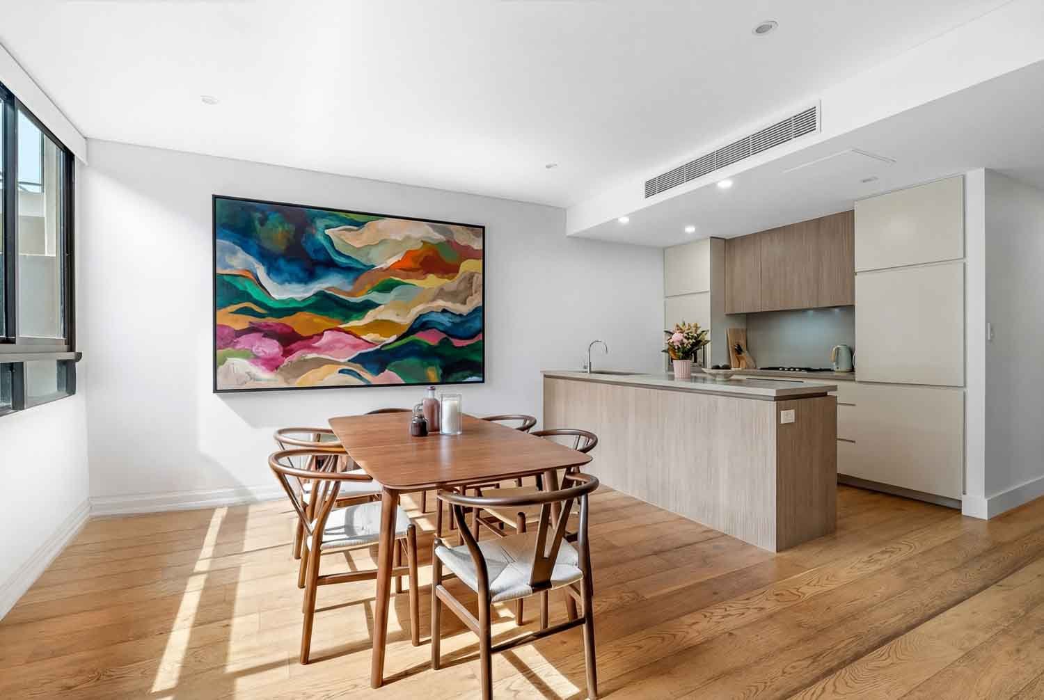 Modern open-concept kitchen and dining area with wooden dining table, six chairs, wall-mounted colorful abstract art, and light wood cabinetry.
