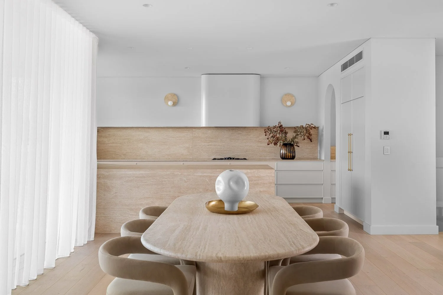 Minimalist dining room with a light wood dining table, six beige chairs, a white decorative vase on a gold tray, and a modern kitchen with beige backsplash and white cabinets in the background.