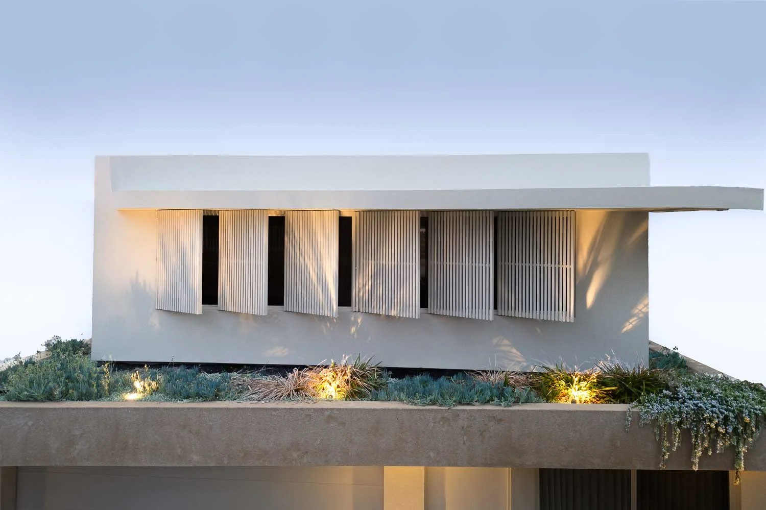 Modern white house with horizontal wooden slats on windows and a rooftop garden illuminated by outdoor lights, under a clear sky.