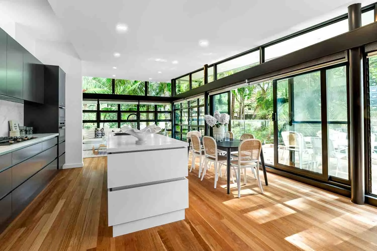 Modern open-plan kitchen and dining area with large glass windows and doors, wooden flooring, white island, black cabinets, and a dining table with chairs and a vase with flowers.