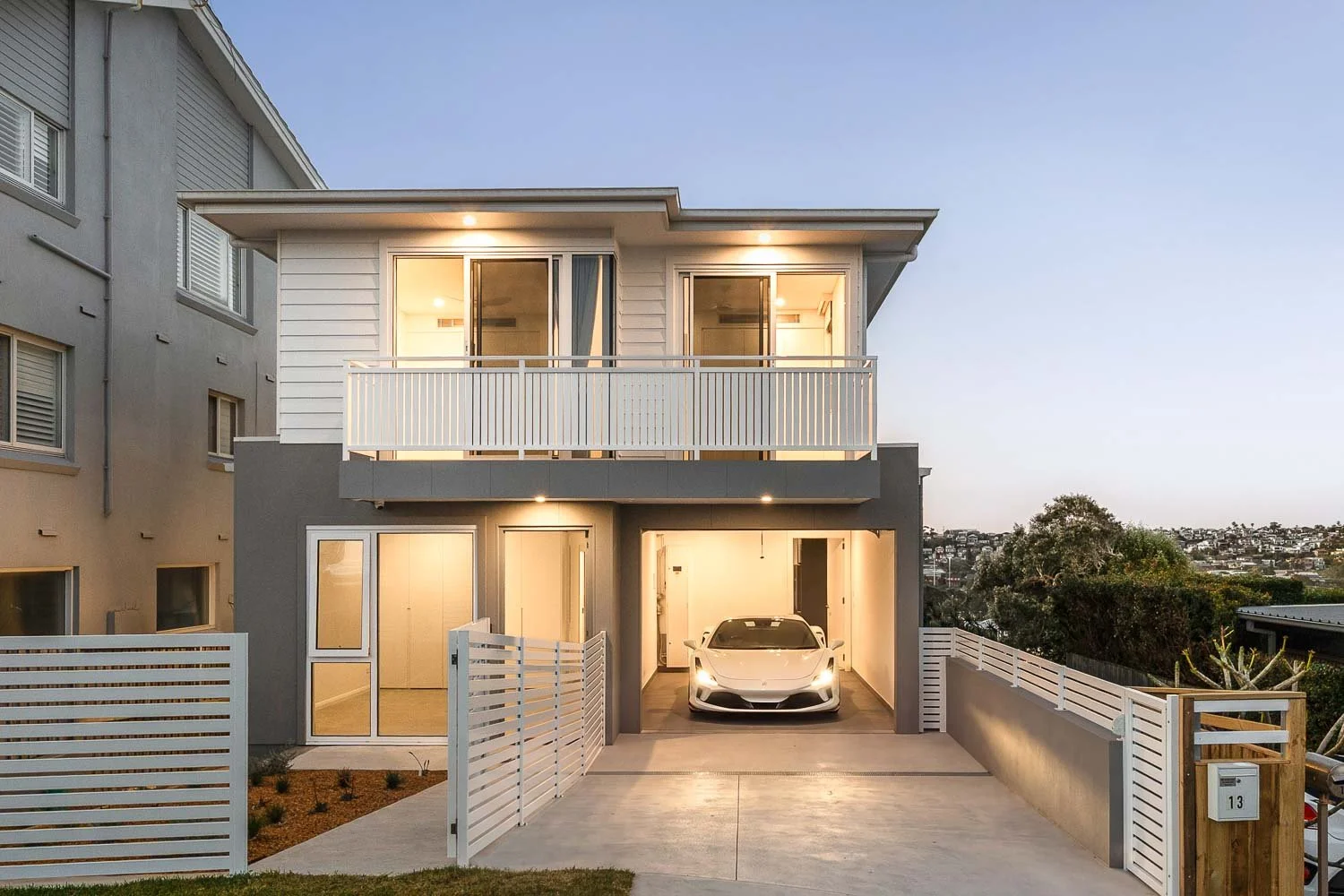 A modern, multi-story residential building with a garage, white fence, and a sleek white sports car parked inside. The building features large windows, a balcony with white railing, and exterior lighting, with a cityscape and trees in the background 