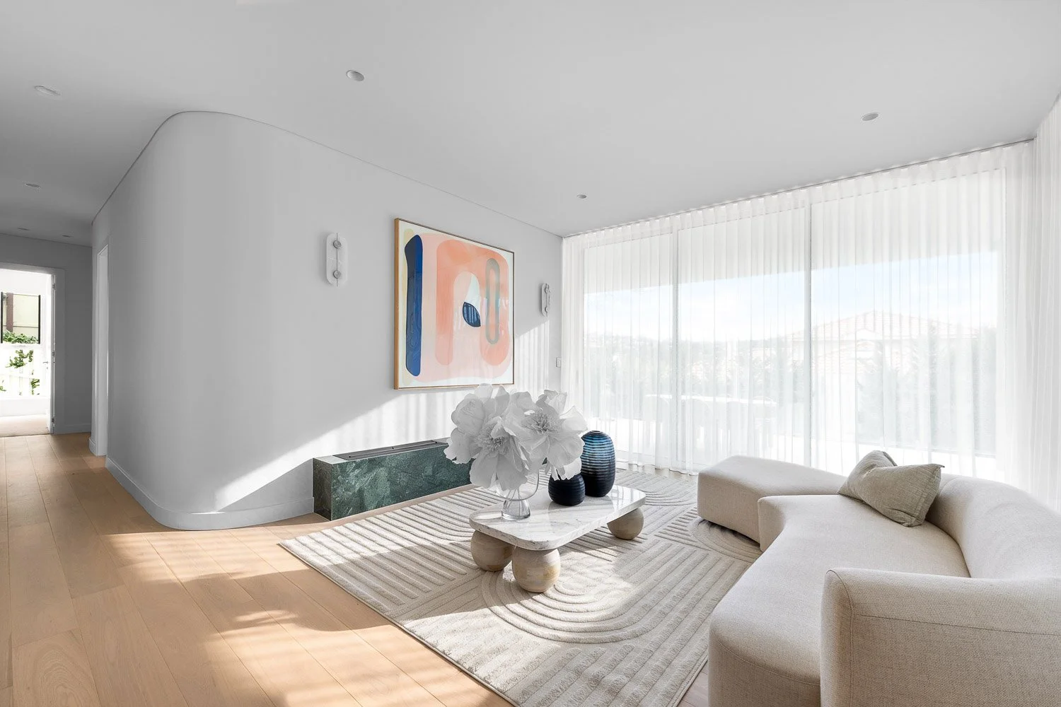 Bright living room with white walls and ceiling, large windows with sheer curtains, modern abstract artwork on the wall, a white curved sofa, a rectangular marble coffee table with decorative vases and flowers, light wood flooring, and a striped area