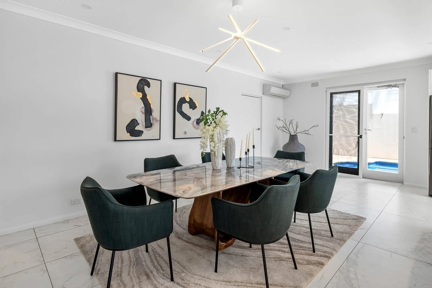 Modern dining room with six dark green chairs around a marble table, white walls, abstract art, decorative vases, and a large light fixture.