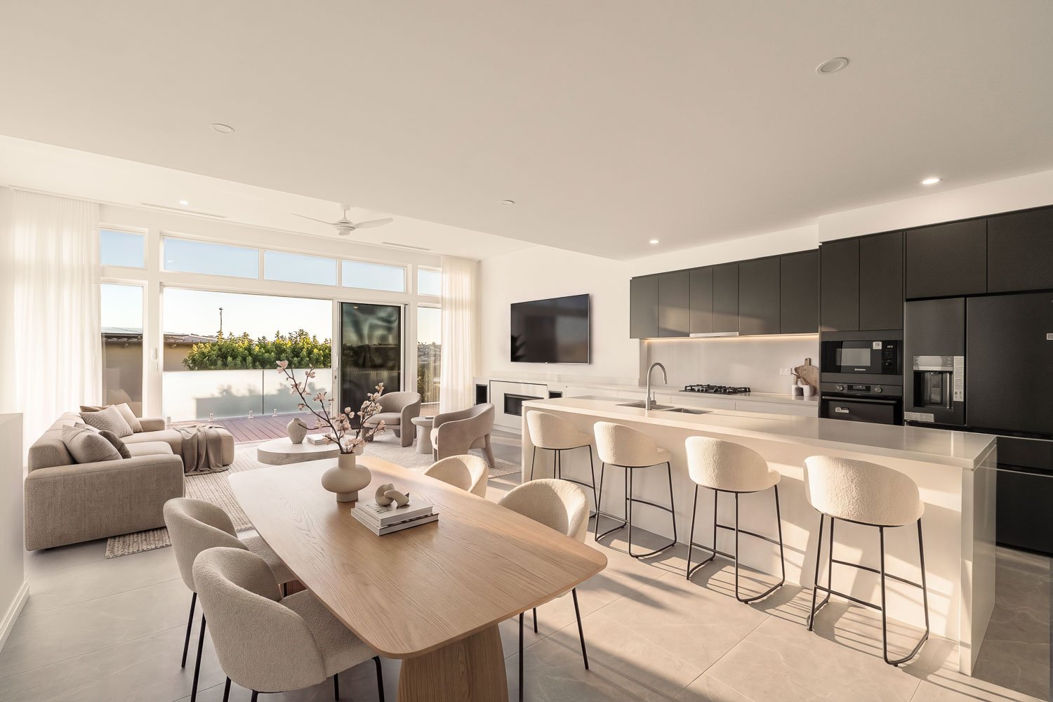 Bright, modern open-concept living room and kitchen with neutral tones, large windows, a dining table, a sofa, a TV, and kitchen appliances.