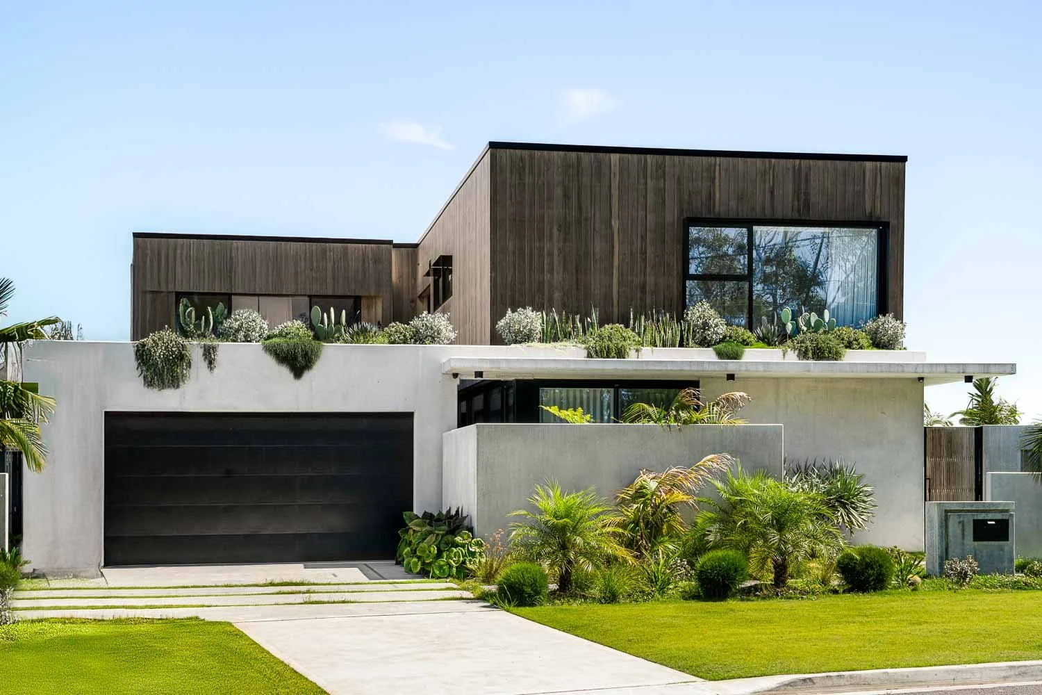 Modern two-story house with concrete and wooden exterior, featuring large windows and landscaped front yard with plants and grass.