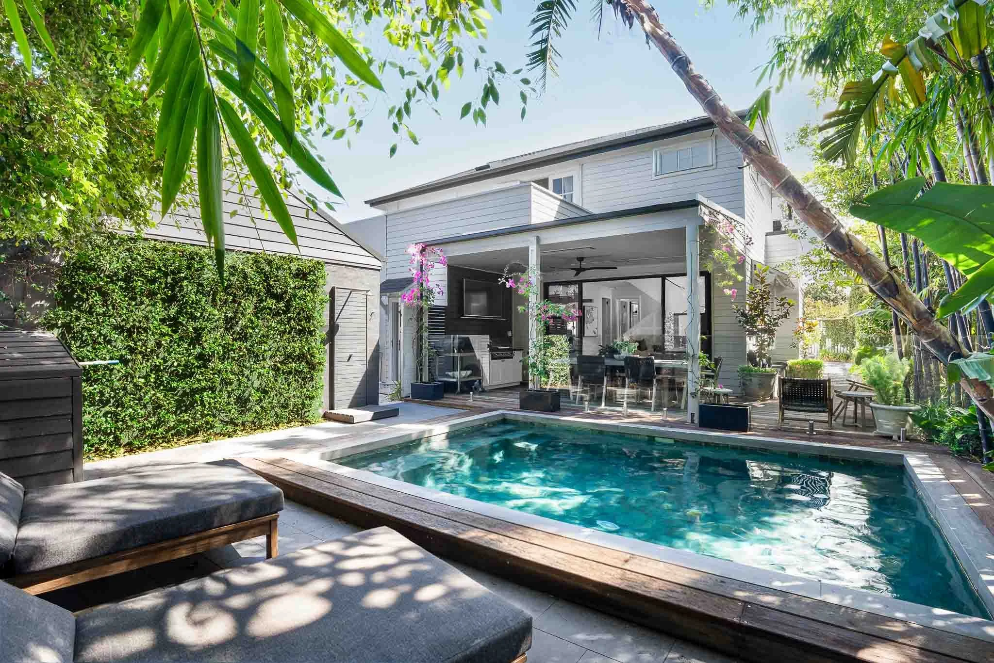 Backyard with swimming pool, outdoor furniture, and a house with a covered patio, surrounded by lush green plants and trees.