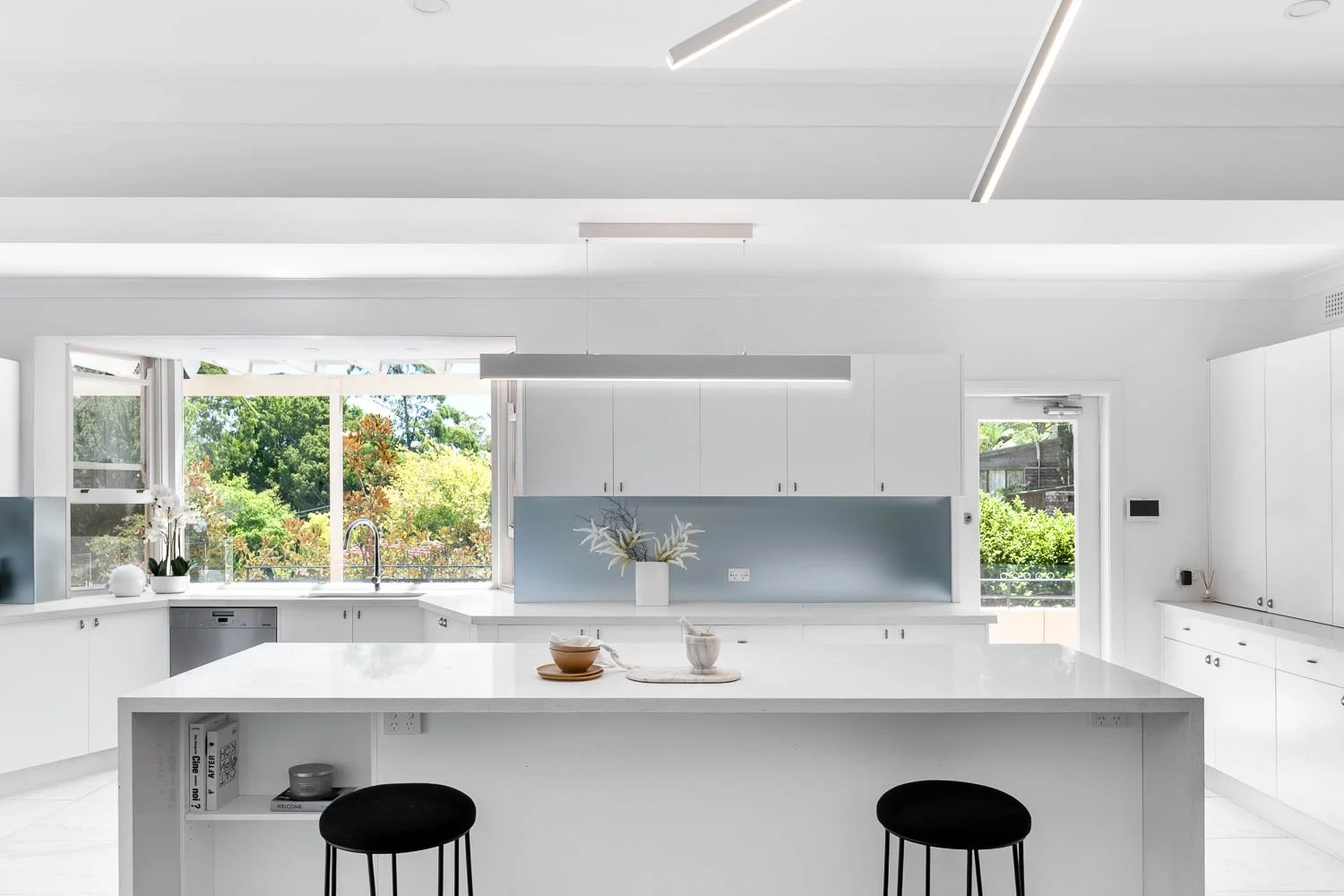 Modern white kitchen with large island, overlooking a garden through open windows, decorated with a vase of flowers and bowls on the island, black stools, and minimalistic cabinets.