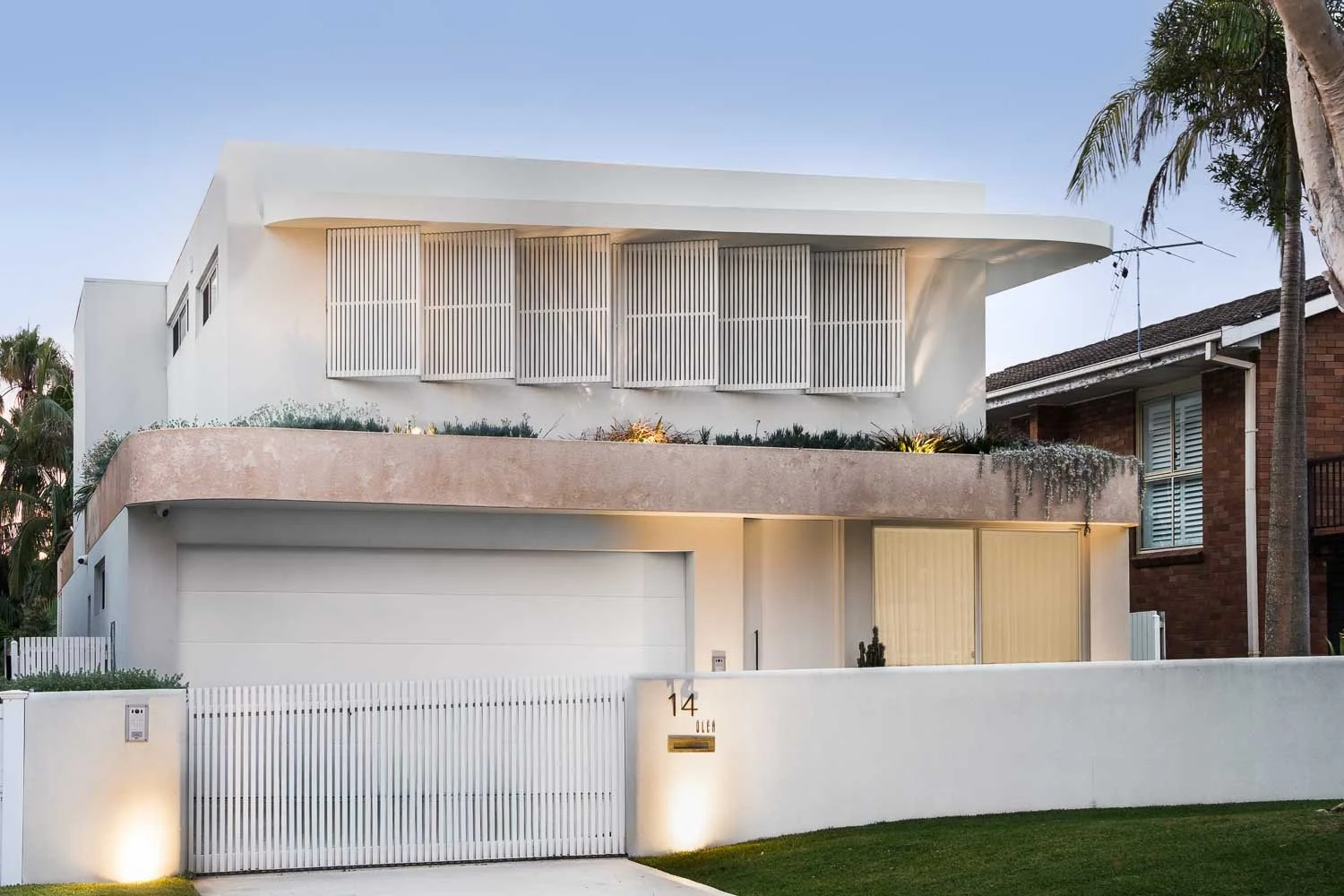 Modern white multi-story house with clean lines, large windows, and a decorated front yard, located in a suburban neighborhood with palm trees.