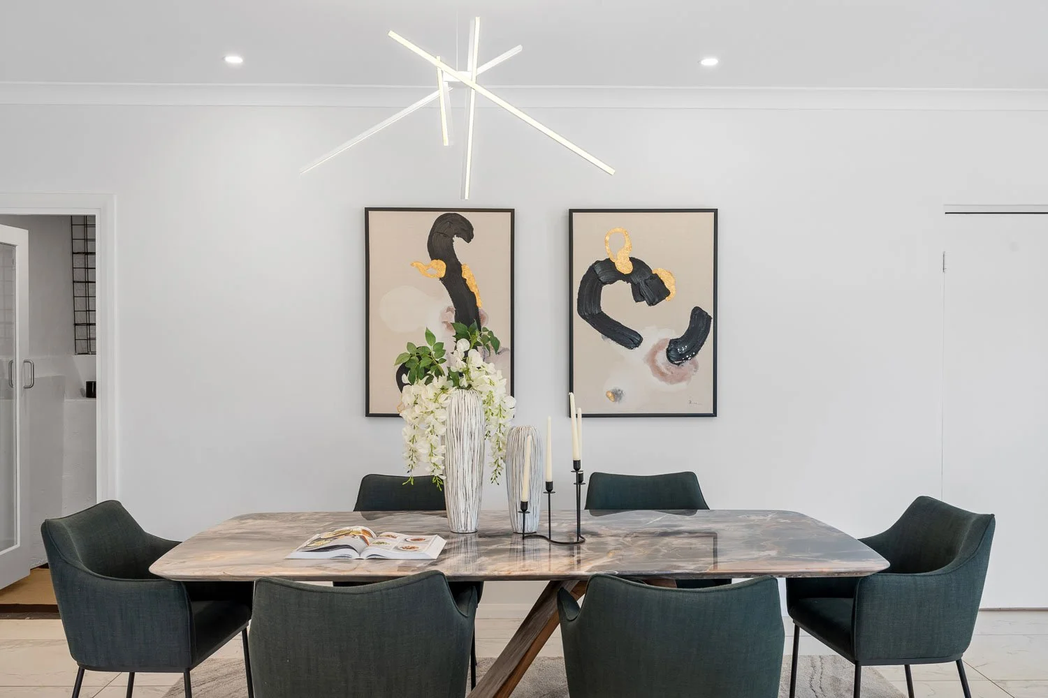 Modern dining room with a polished marble table, six dark green chairs, white walls, two abstract artwork pieces on the wall, and a tall white flower arrangement on the table.