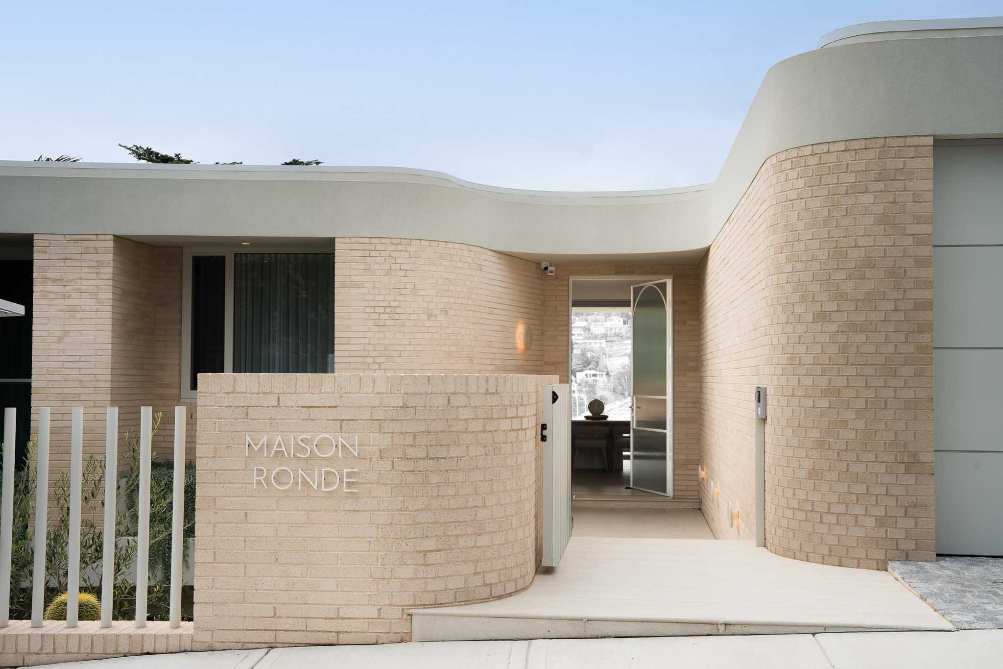 Front entrance of a modern house with brick walls, a curved wall with the sign 'MAISON RONDE,' metal gate, and open door showing a view inside.
