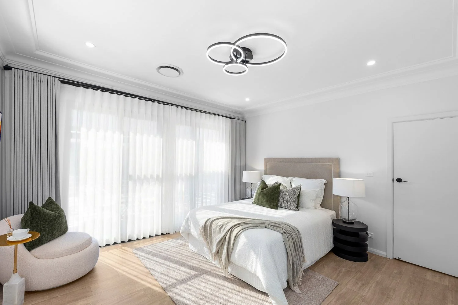 Modern bedroom with white bedding, beige headboard, green and gray pillows, nightstand with white lamps, large window with sheer white curtains, light wood floor, and a curved cream chair with green pillow and small table.
