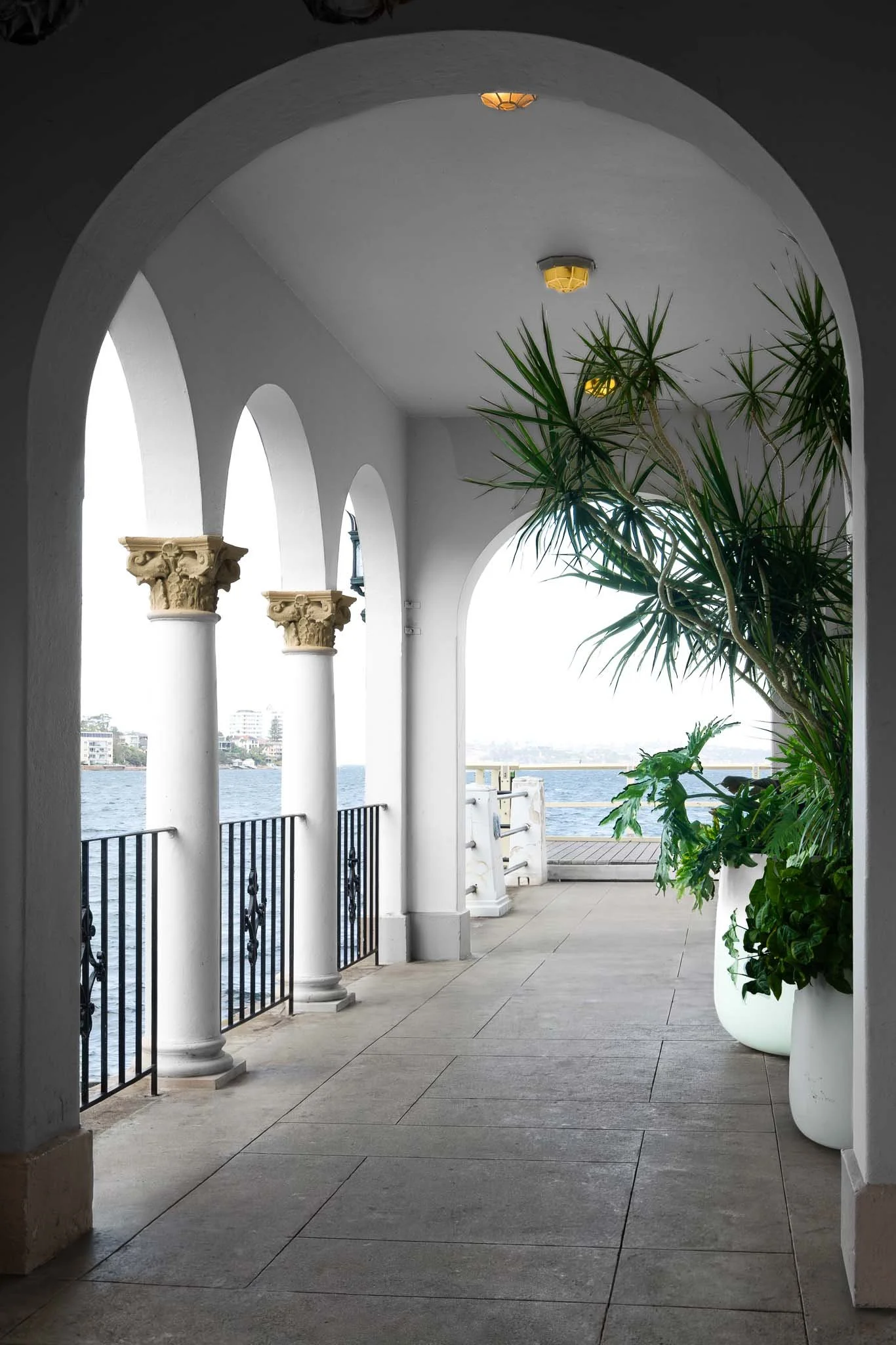 View of a courtyard with white arches, columns with ornate capitals, potted plants, and a waterfront view in the background.