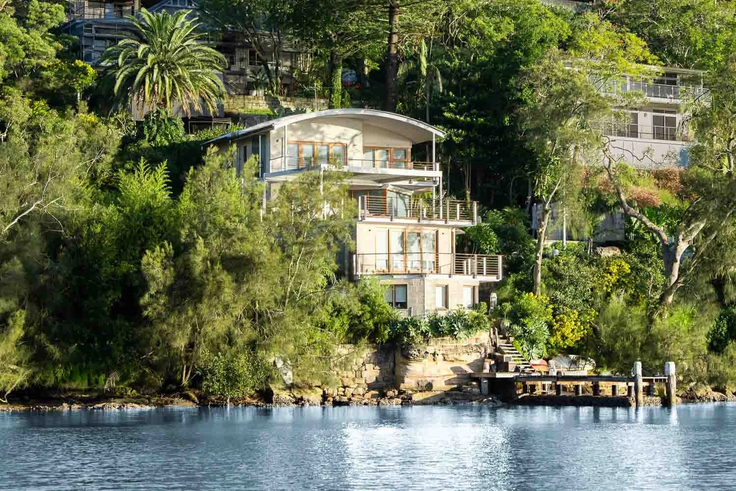 A modern house built on a hillside surrounded by dense green trees, with a body of water in the foreground and a small dock extending over the water.