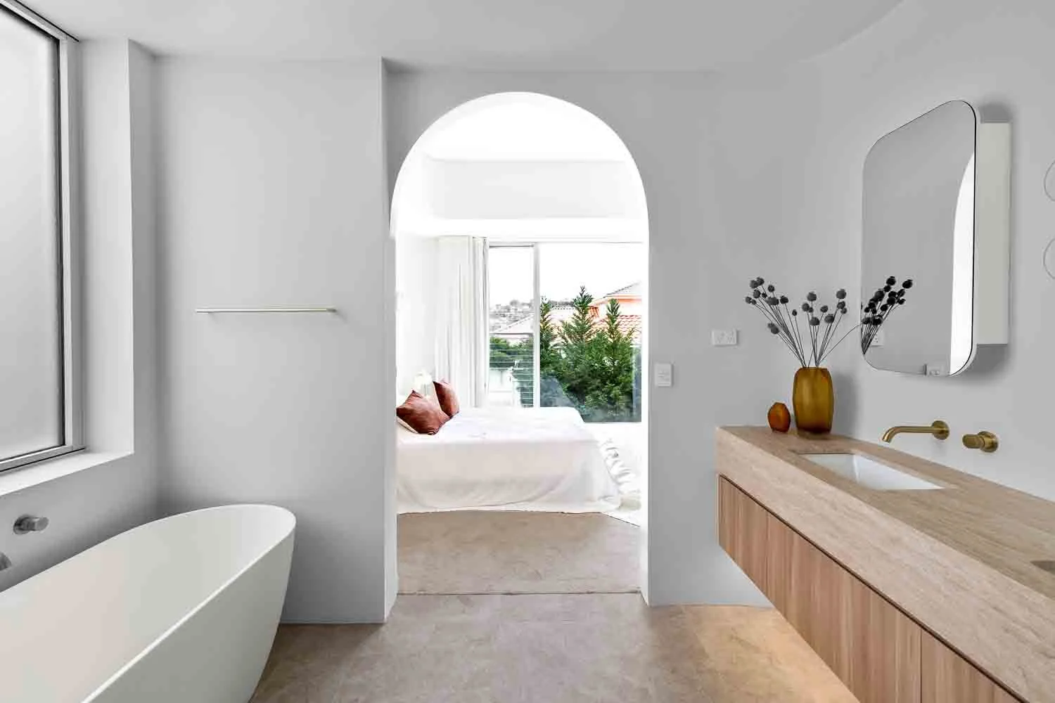 Modern bathroom with a free-standing bathtub on the left, a vanity with a mirror and two vases on the right, and a doorway leading to a sunlit bedroom with a large window and bed with cushions in the background.