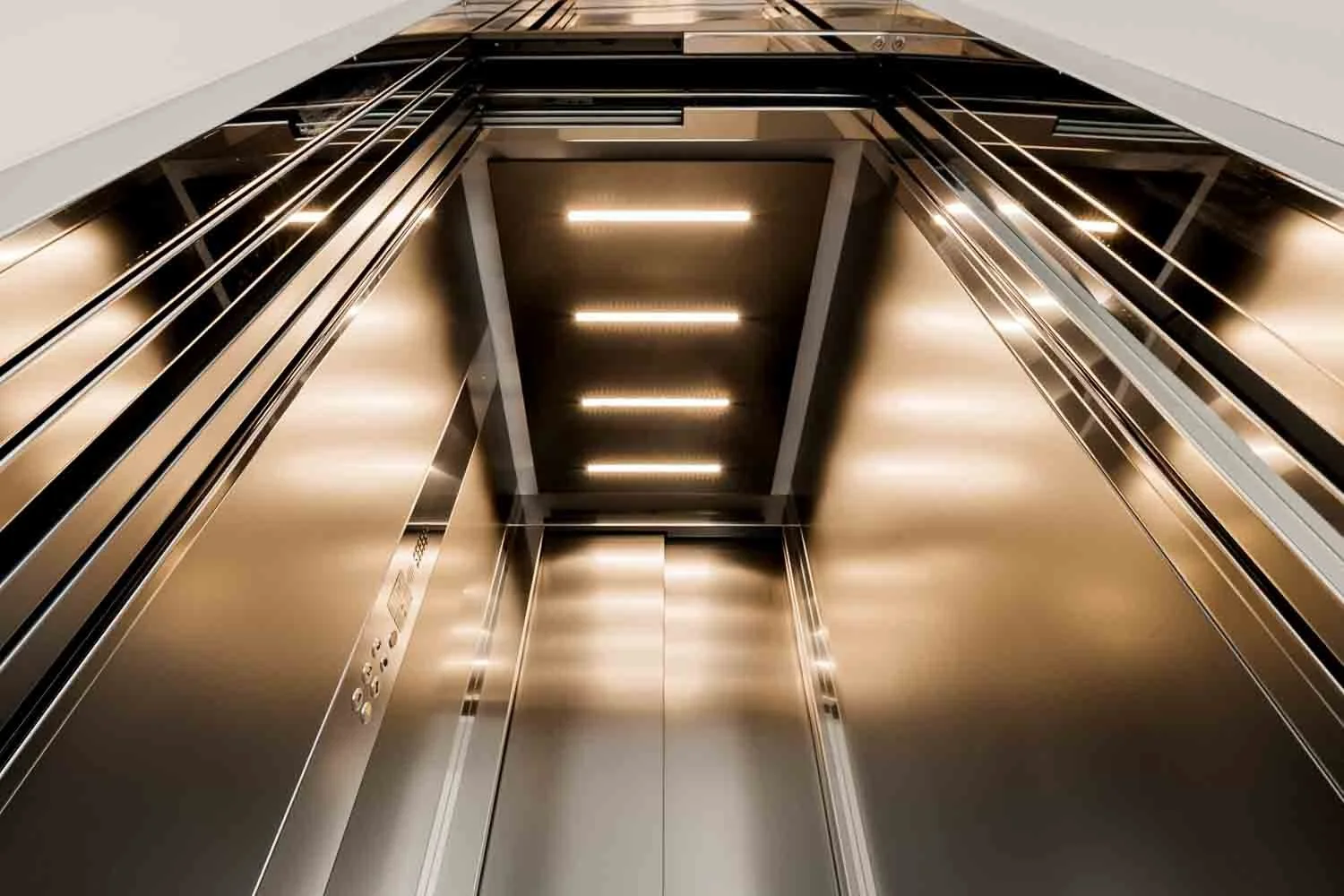 Inside of a modern, empty elevator with metallic walls and ceiling, looking upward from the floor level to the ceiling, with bright lighting.