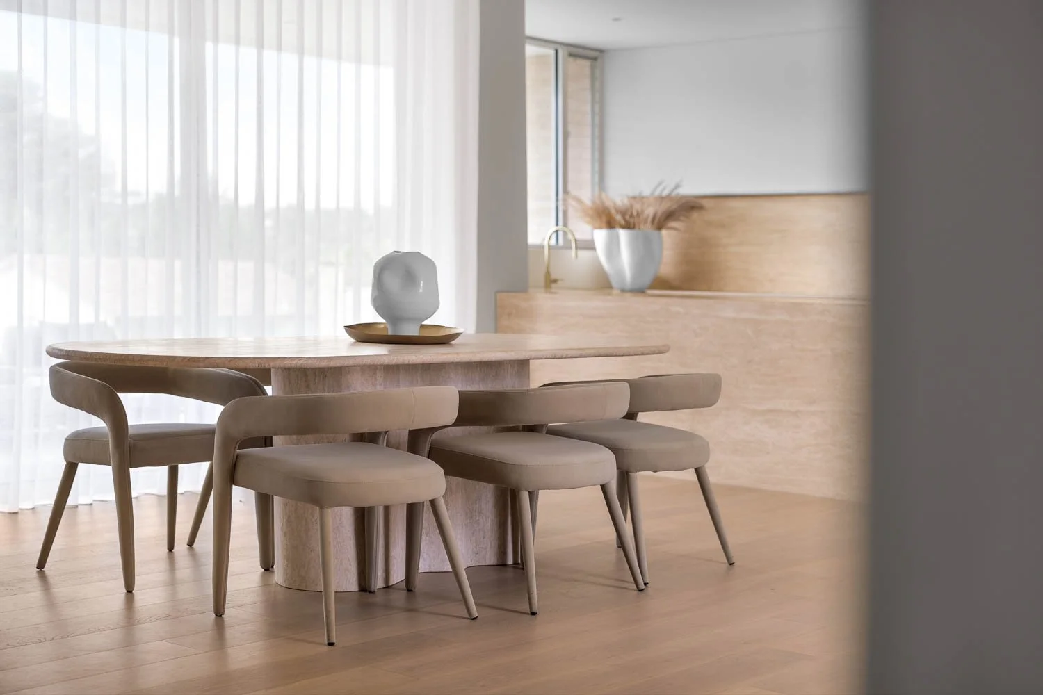 Modern dining room with a light wood table and matching chairs, a decorative ceramic vase on the table, and a large white vase with dried pampas grass on a ledge near a window with sheer curtains.
