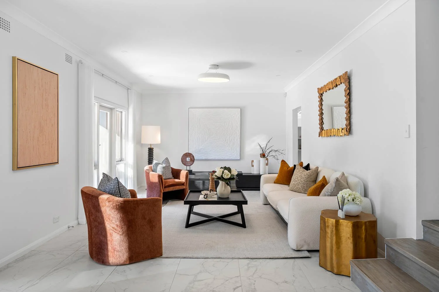Living room with white walls, marble floor, and light-colored furniture. Contains a white sofa with multicolored pillows, red armchairs, a black coffee table with a flower vase, decorative vases and artwork on the walls, and a wooden side table with 