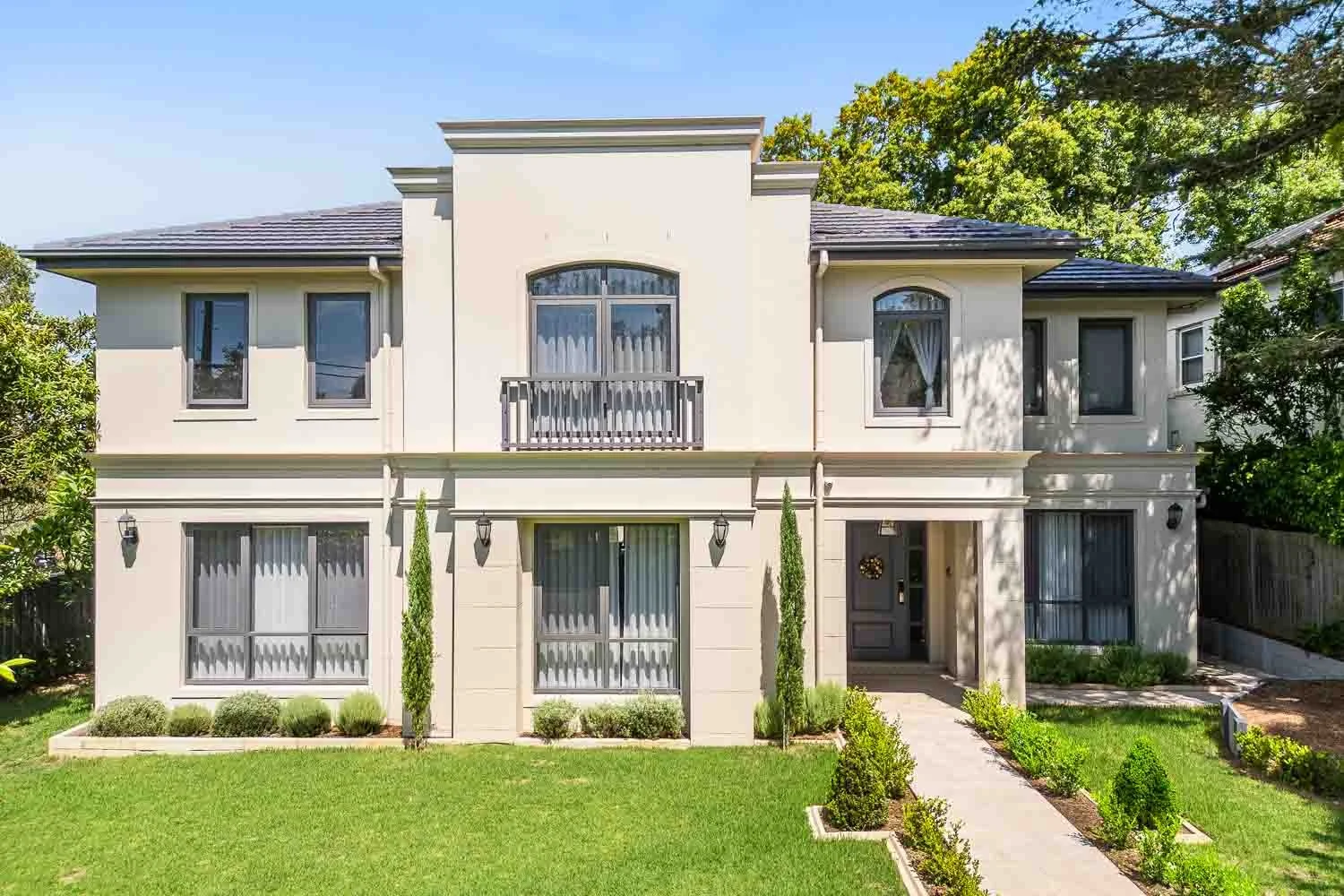 A modern, two-story house with a white exterior, multiple large windows with curtains, and a small balcony on the upper floor. The front yard features a well-maintained lawn, small bushes, and a pathway leading to the front door. Tall, narrow trees a