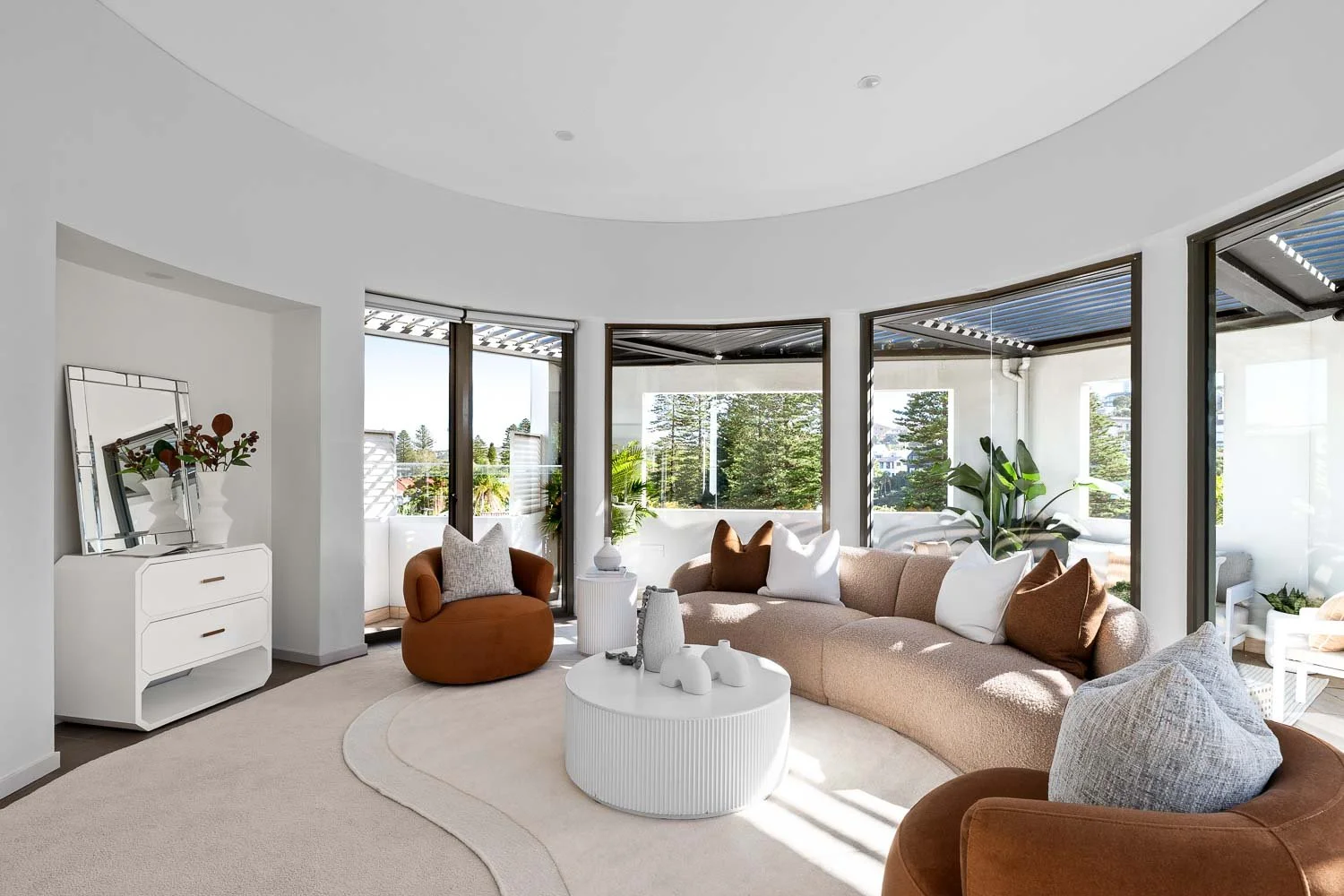 Bright modern living room with beige sectional sofa, brown and white pillows, round white coffee table with decorative vases, large windows with outdoor views, and green plants.