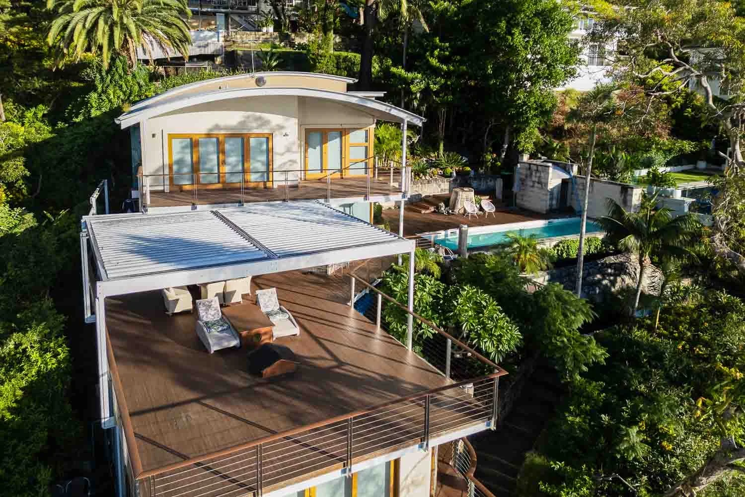Aerial view of a modern house with multiple wooden decks, outdoor seating, a pool, lush green trees, and surrounding foliage.