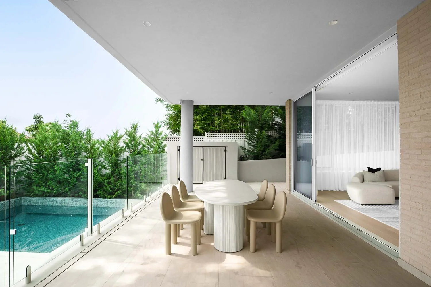 Modern patio with glass railing, outdoor dining table with six beige chairs, swimming pool, and view into an indoor living room with white sofa and sheer curtains.