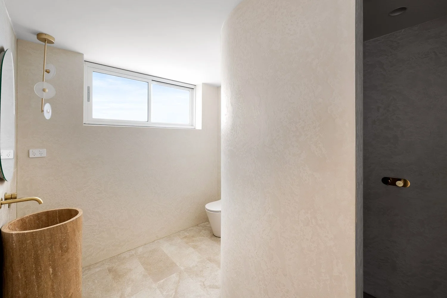 Modern bathroom with beige textured walls, a small window, a gold and white light fixture, a beige stone sink, a toilet, and part of a dark shower area.