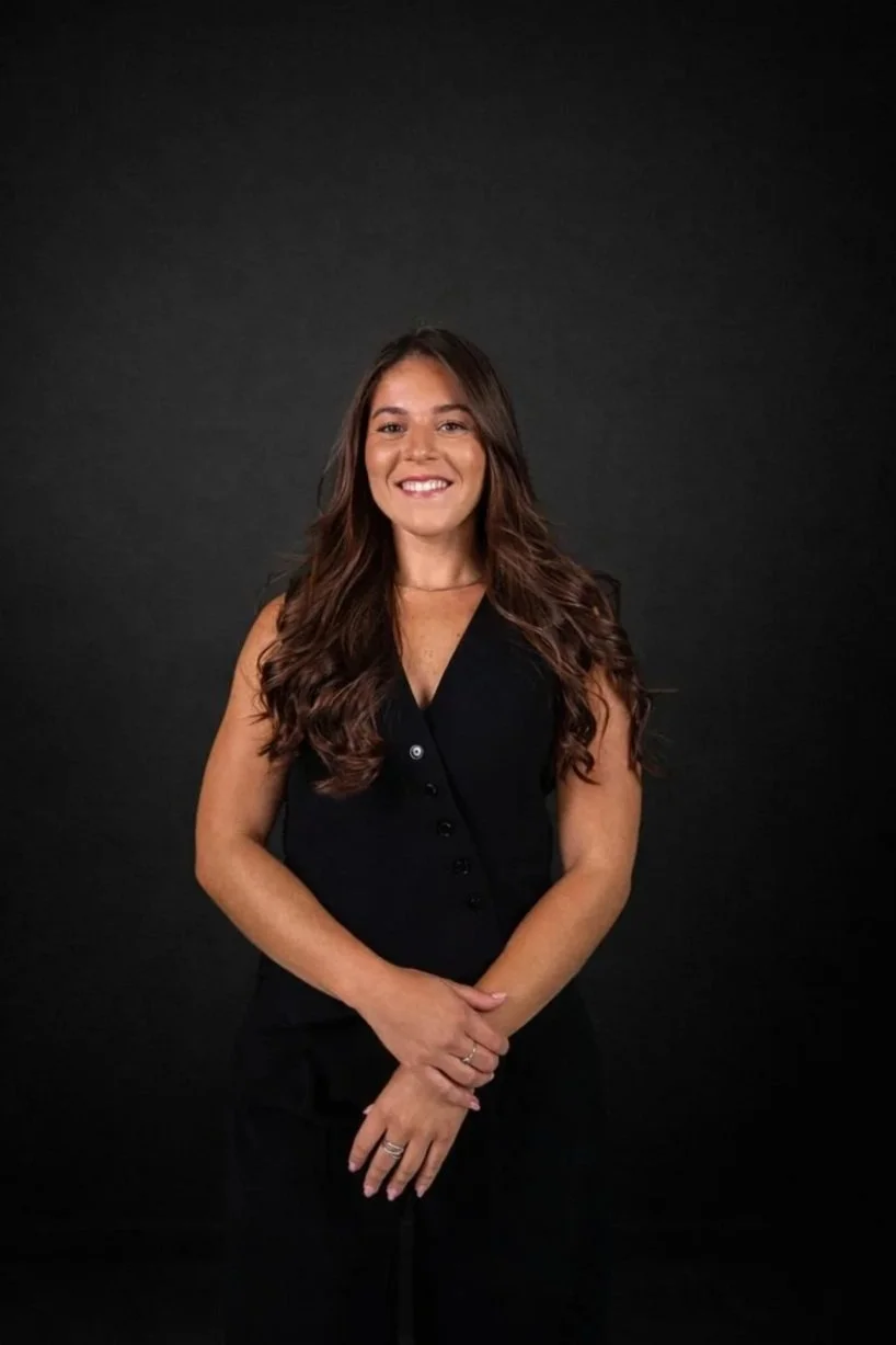 A woman with long dark hair, light skin with freckles, wearing a blue blouse and a checkered blue and white blazer, smiling against a wooden background.