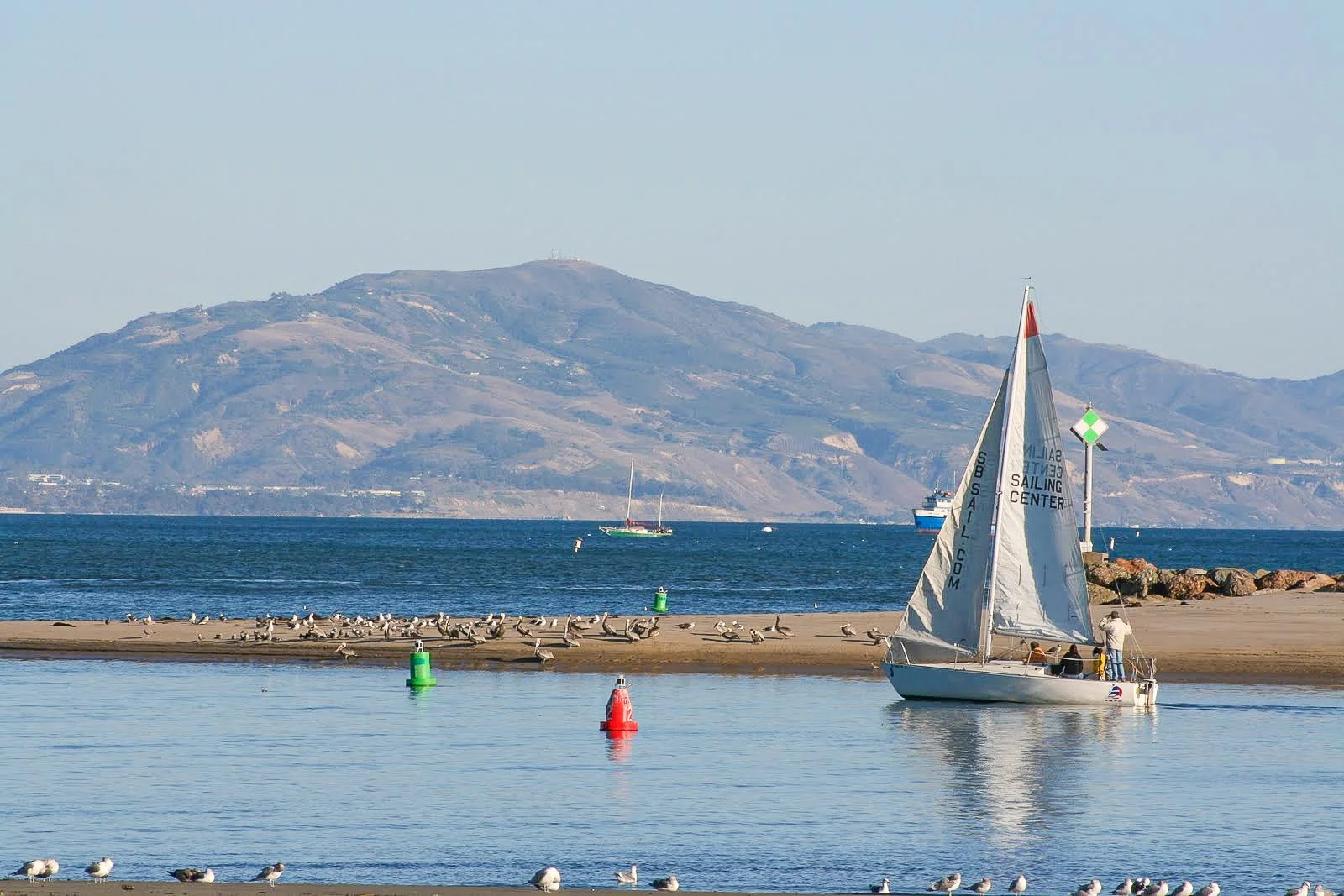 Sailboat near the shore with birds on a sandbar and mountains in the background.