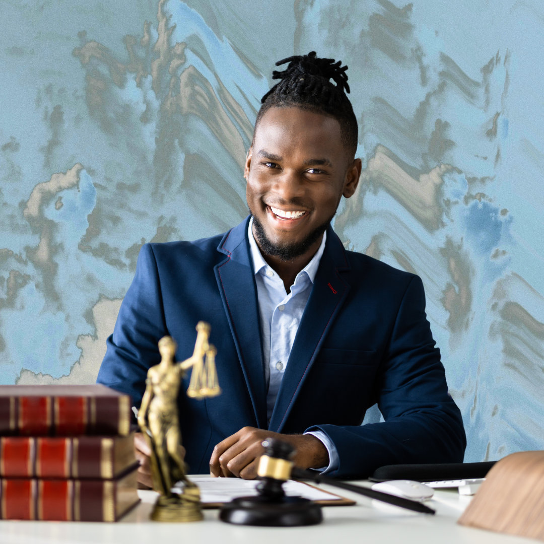 A Black person in a navy blue suit sits at a table in front of paperwork. They sit in front of a marbled blue background.