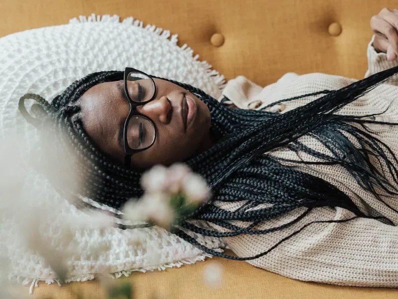 a black person with long braids and a white sweater lays on a yellow couch. their head rests on a circular white cushion.