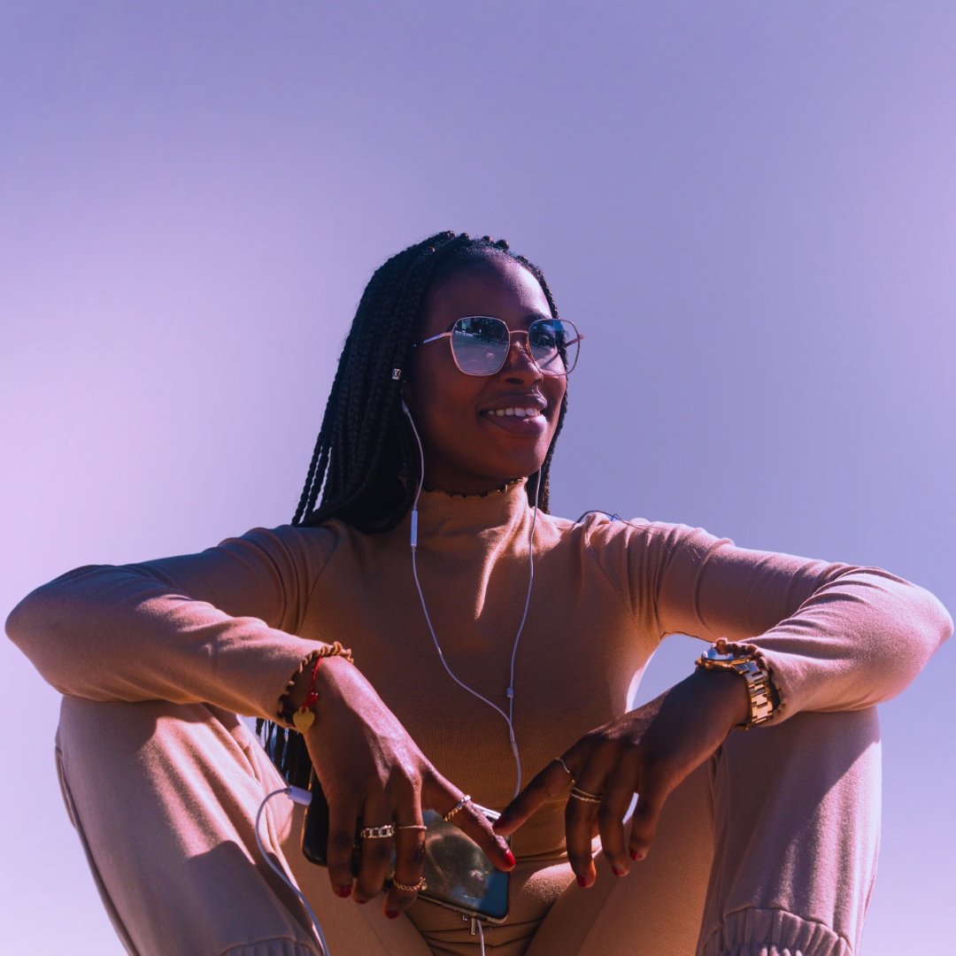 A Black person with sunglasses and wired headphones sits smiling with their arms resting on their knees. They wear a pink short and pants and are in front of a lavender and pink background.
