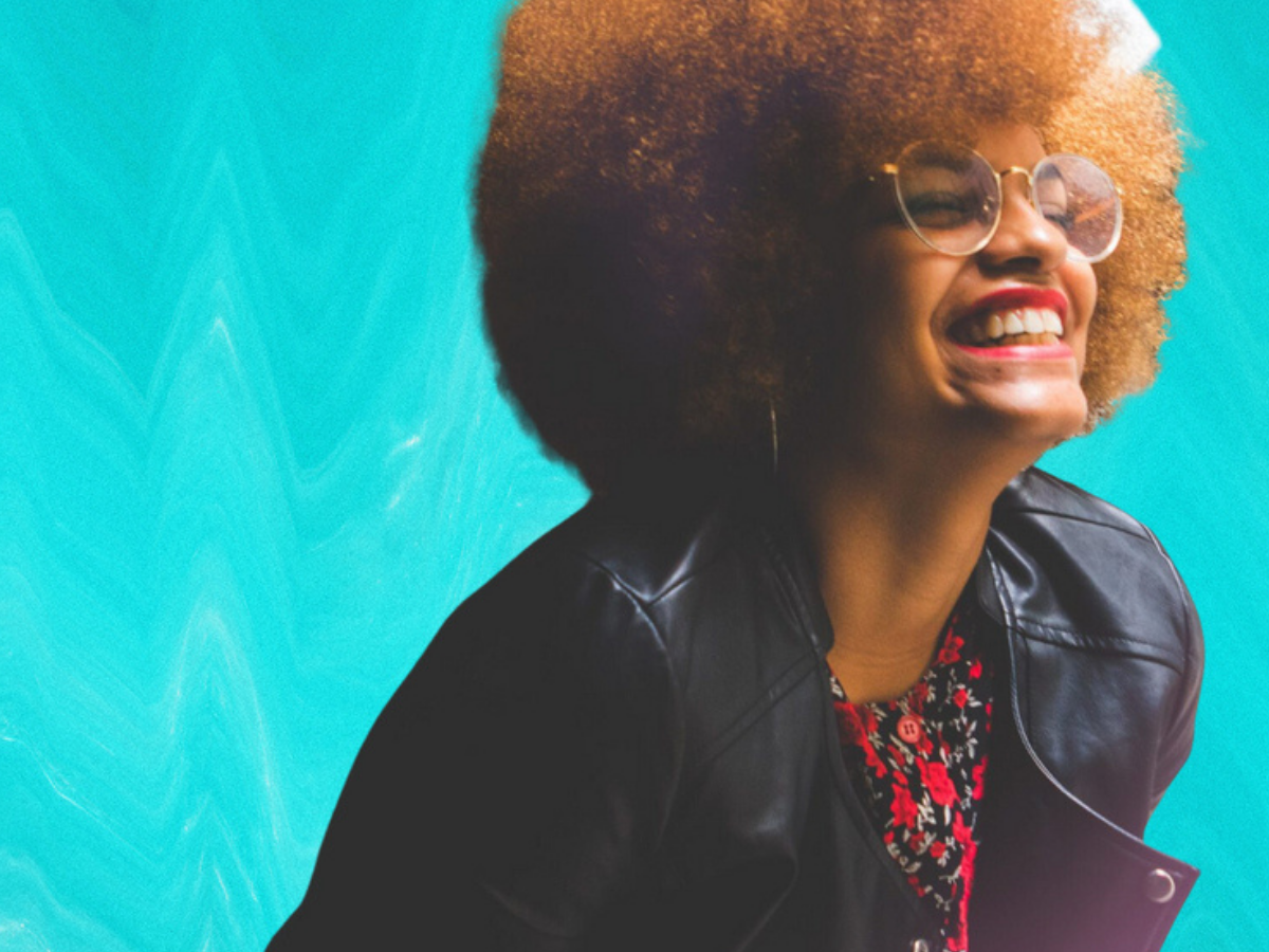 A Black person with a ginger-colored Afro laughs in front of a teal blue background.