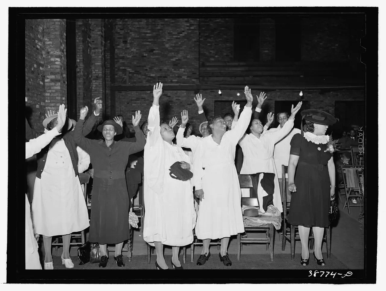 a group of black people raise their hands in praise. some carry hats, and some wear black dresses or skirts while others wear white.