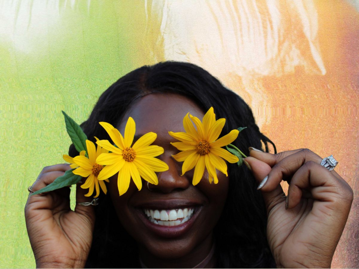 A Black person smiles while holding yellow flowers over their eyes. They are in front of a green and orange background.
