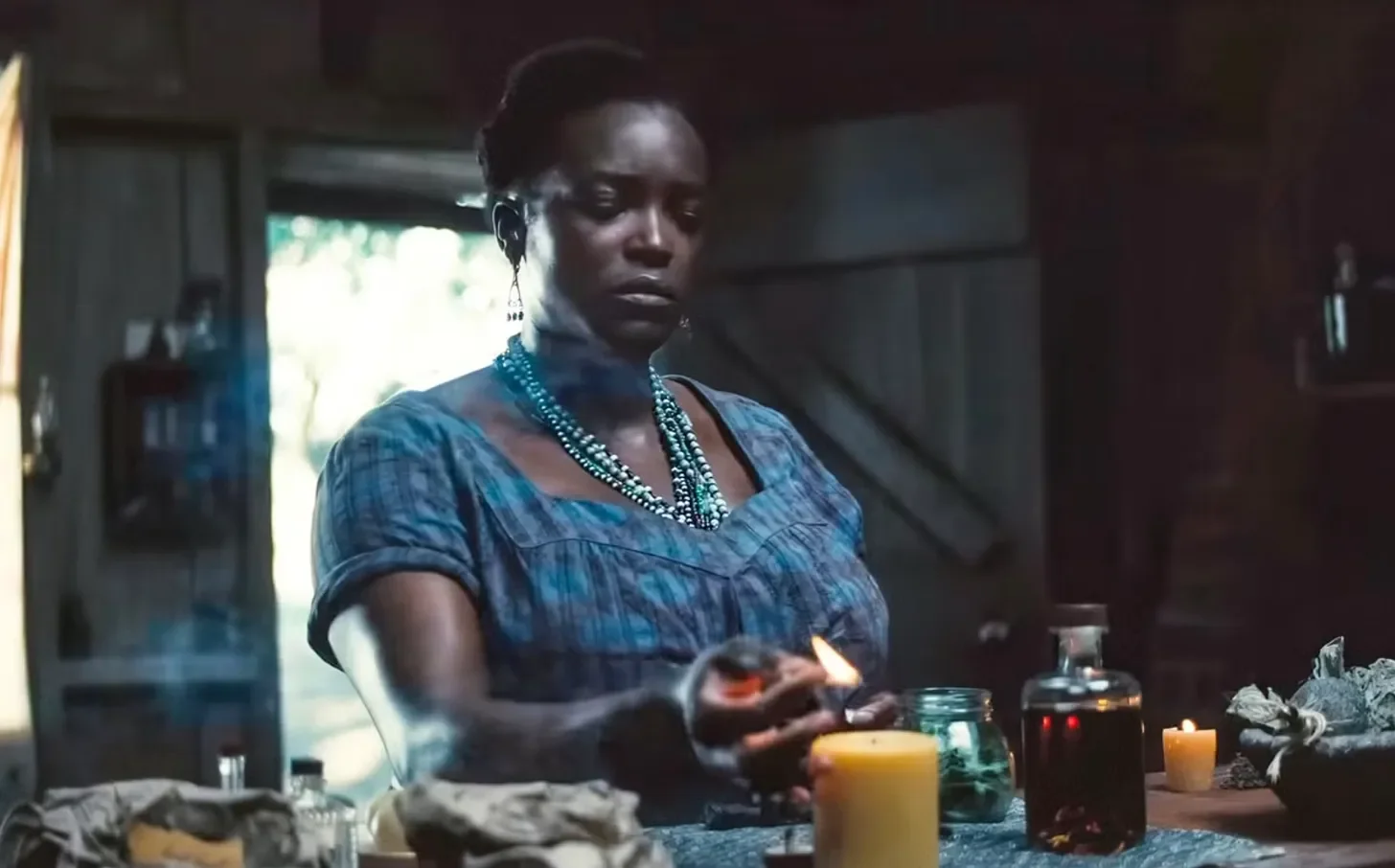 stil from the movie sinners, showing annie, a black woman in a striped blue dress and layered beaded necklaces, lighting a candle on a tabletop in front of her.